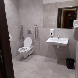 Firma Wykończeniowa Sebastian Tokarczyk - Jasna łazienka dla niepełnosprawnych z toaletą z uchwytami, umywalką i lustrem, w którym odbija się osoba robiąca zdjęcie.