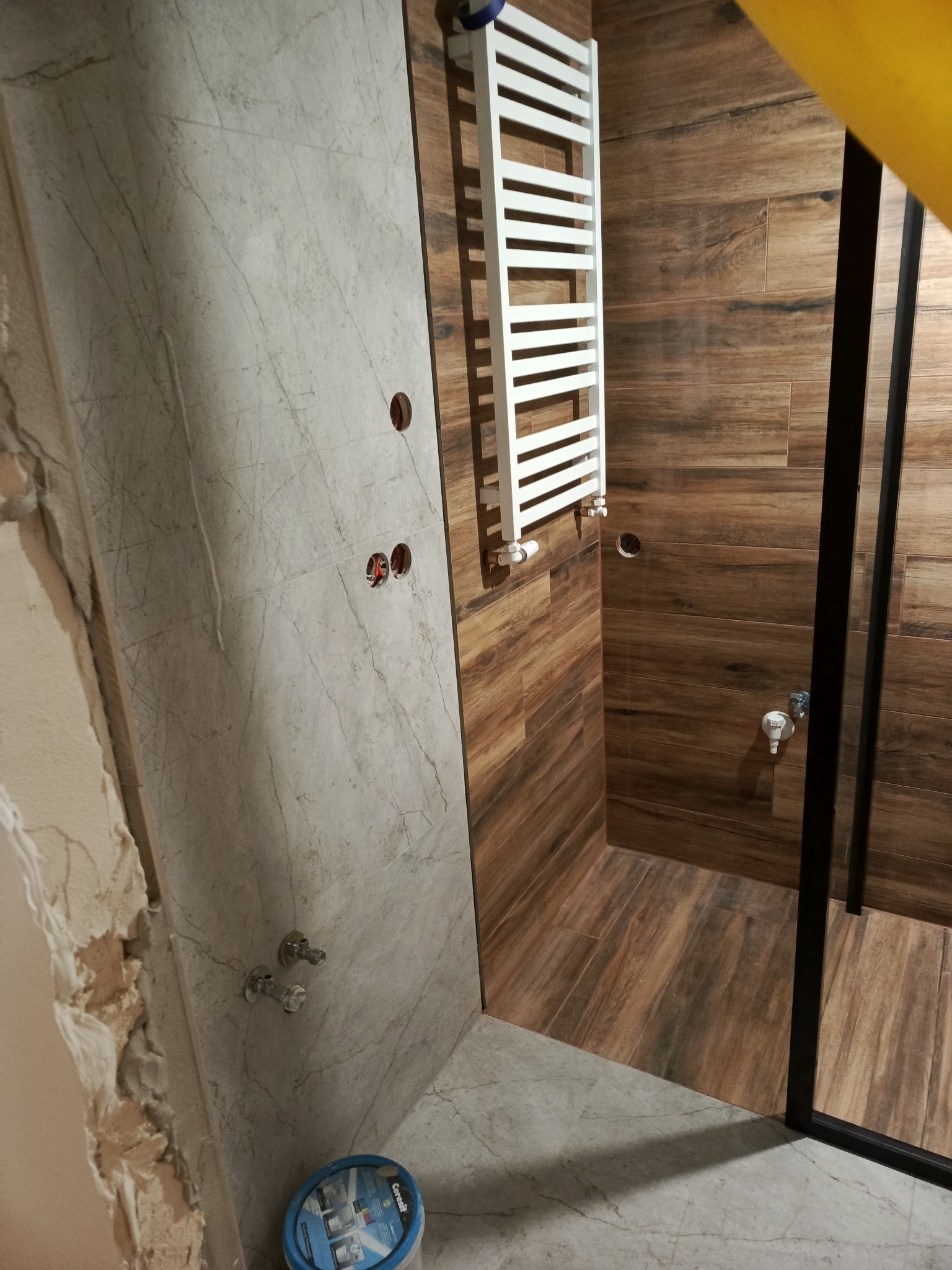 Łazienka w trakcie wykańczania: płytki imitujące drewno i marmur, grzejnik drabinkowy, kabina prysznicowa z czarnymi profilami. Widoczne przygotowane punkty instalacji.