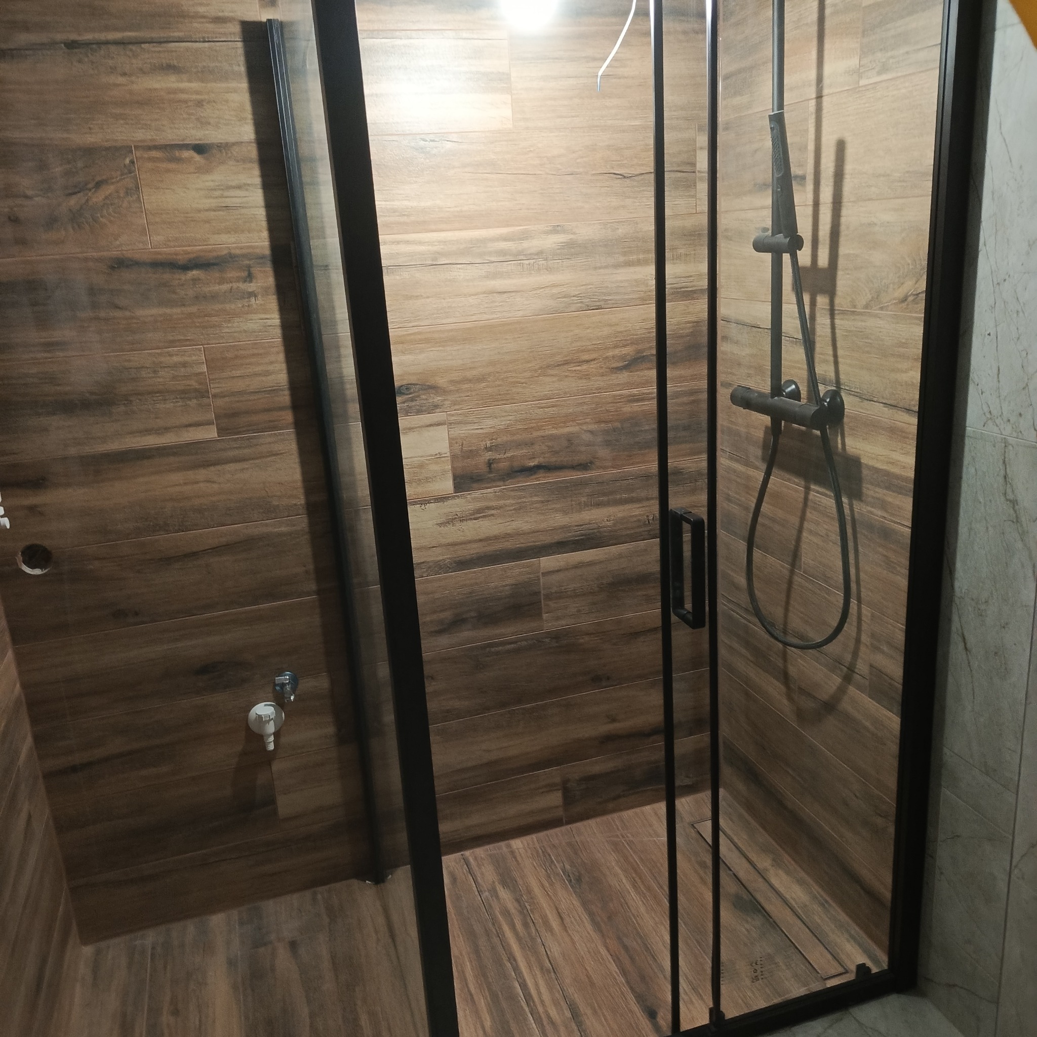 Kabina prysznicowa z czarnymi profilami, wykończona płytkami imitującymi drewno. Widoczna bateria i odpływ liniowy. Minimalistyczny design.