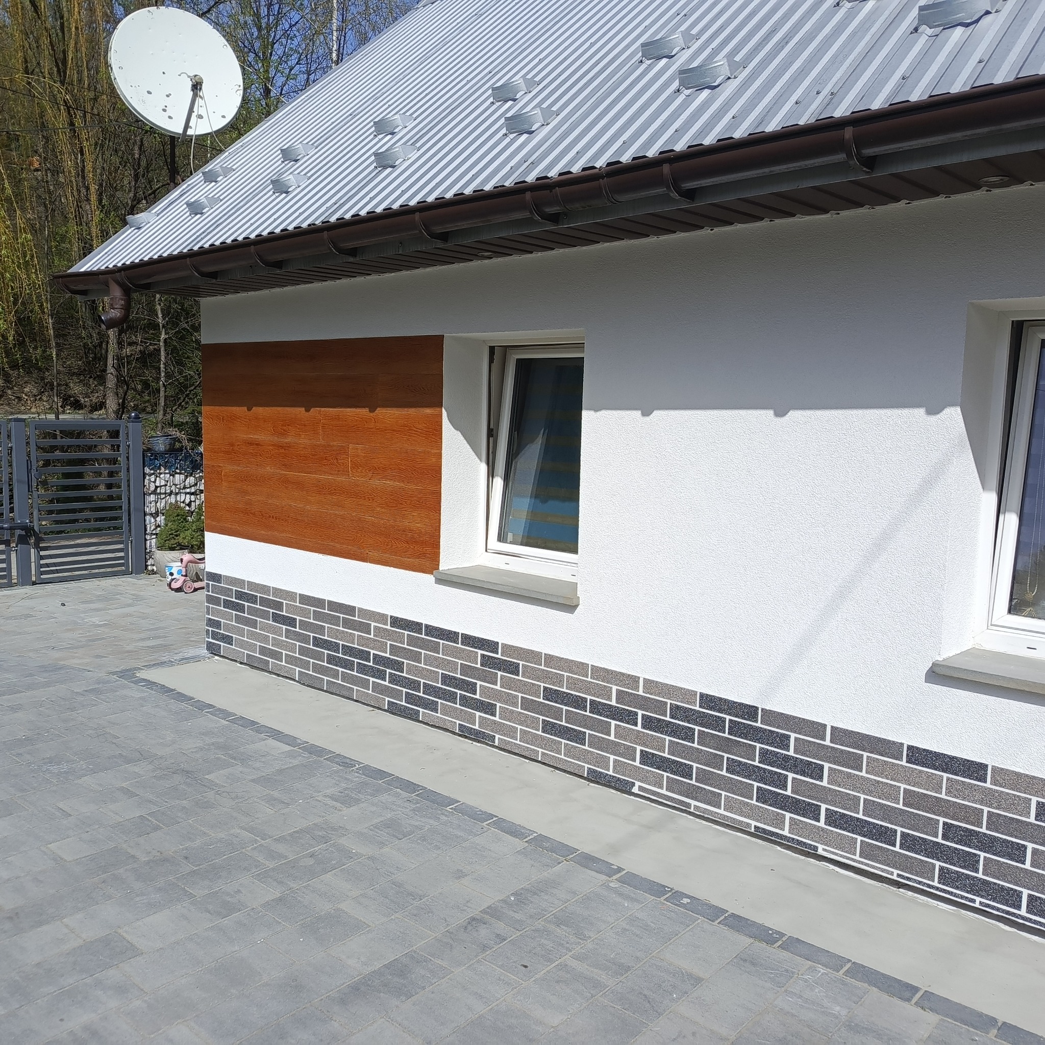 Elewacja domu z elementami dekoracyjnymi: panel drewnopodobny, cokół z cegły klinkierowej, biały tynk strukturalny. Na dachu widoczna antena satelitarna.