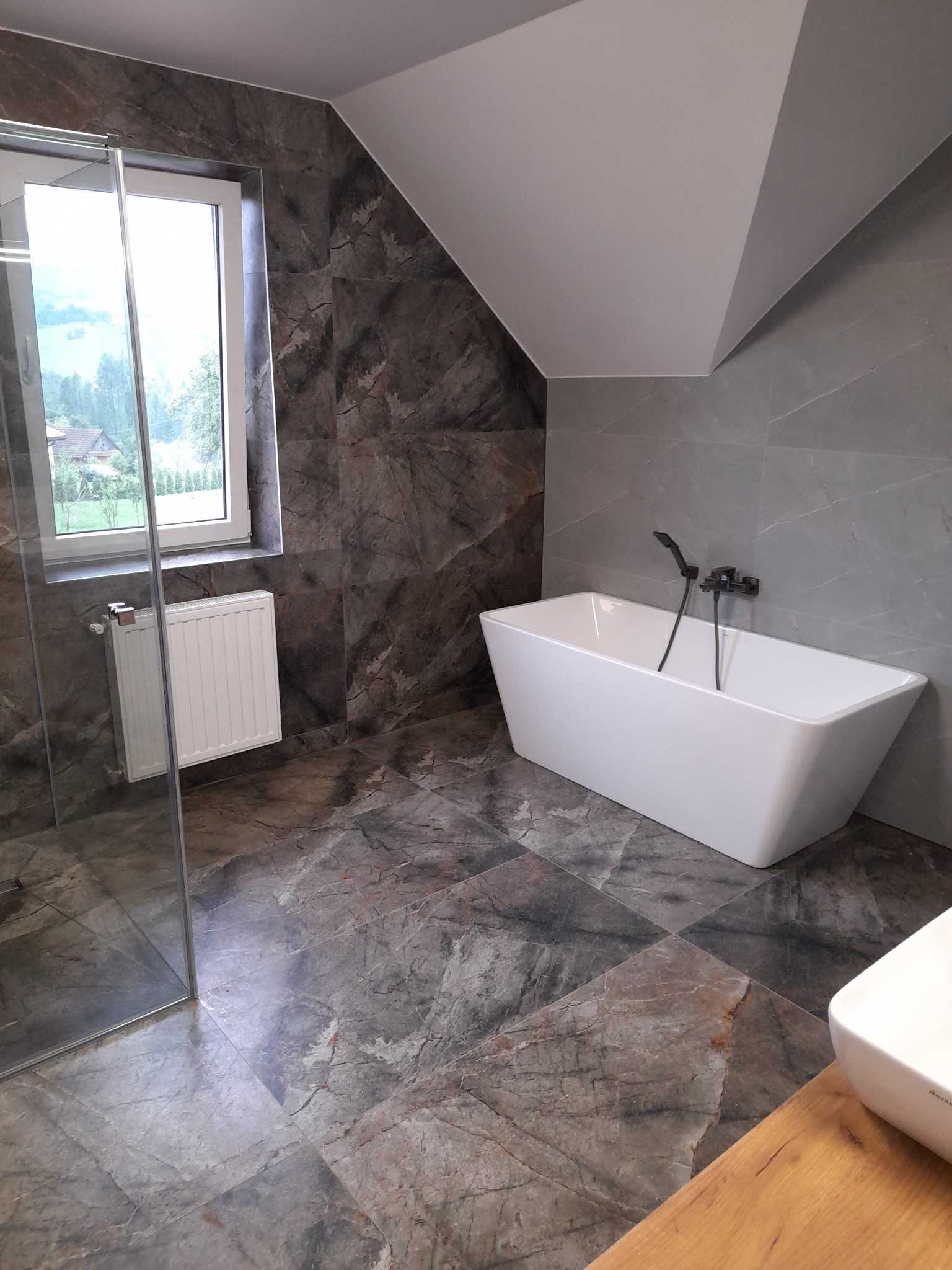 Nowoczesna łazienka na poddaszu z marmurową glazurą, białą wanną wolnostojącą i grzejnikiem pod oknem. Kabina prysznicowa ze szklanymi drzwiami.