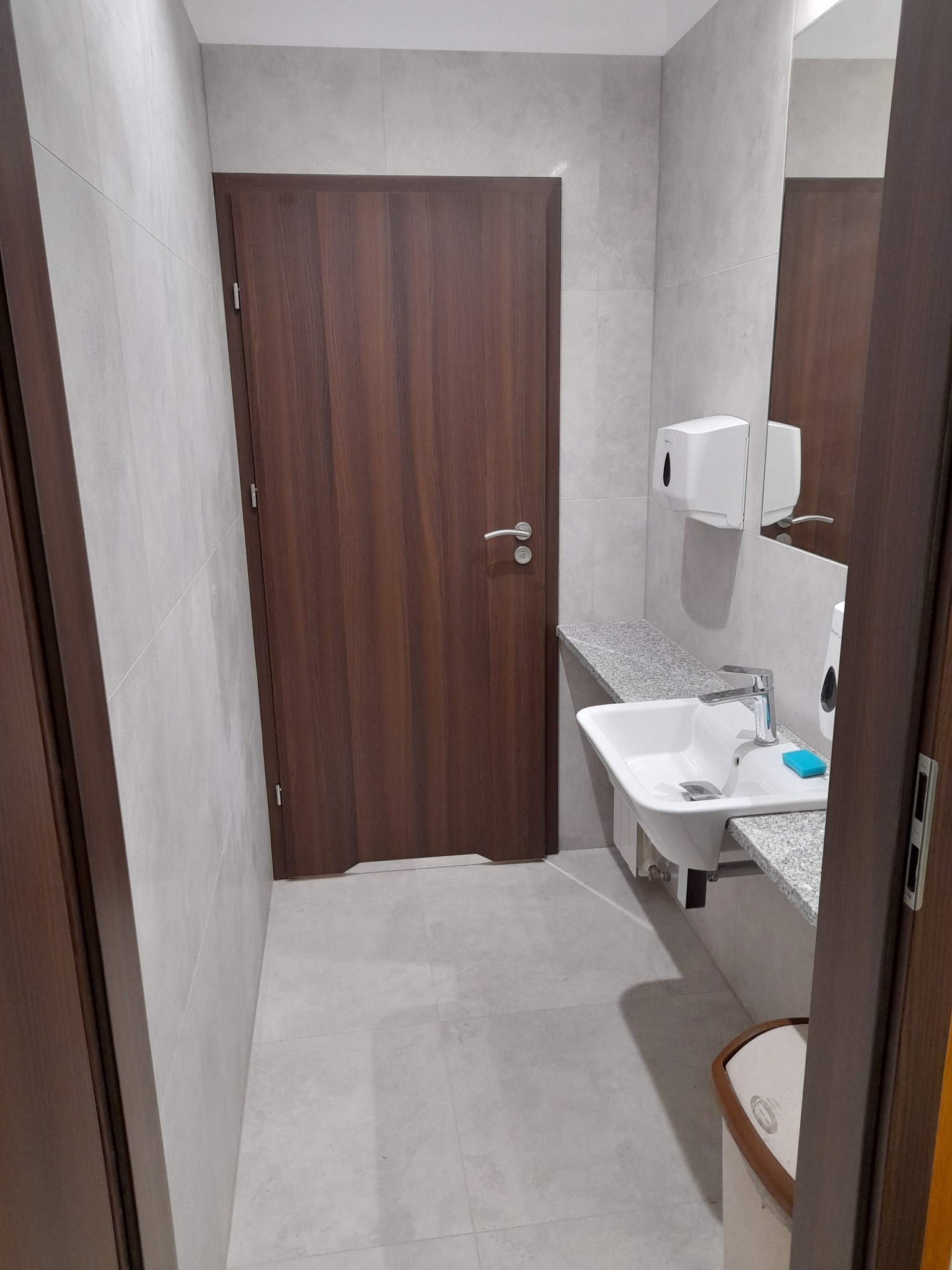 Nowoczesna łazienka z umywalką nablatową na szarym blacie, brązowe drzwi i jasne płytki tworzą minimalistyczną przestrzeń. Widoczny dozownik mydła i lustro.