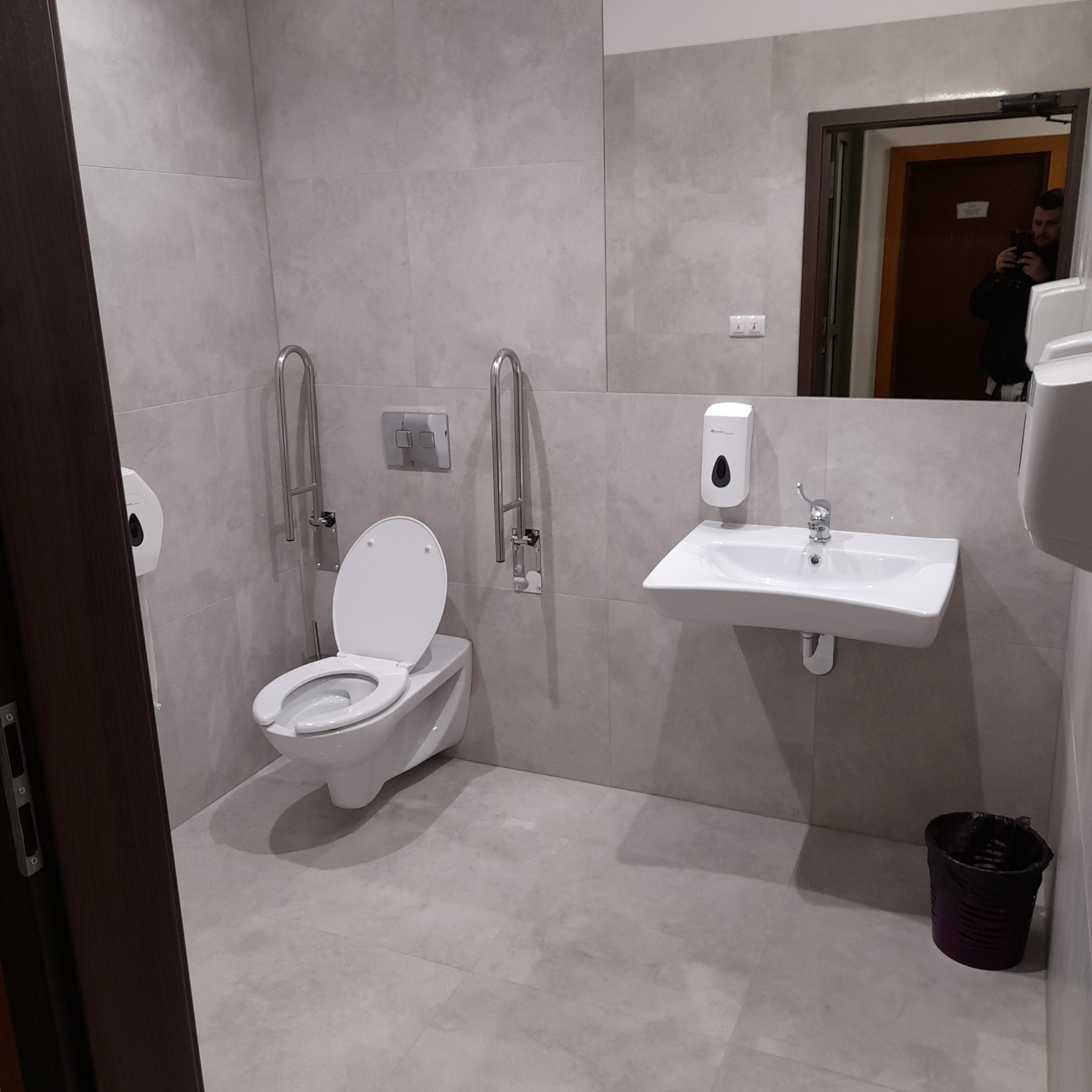 Jasna łazienka dla niepełnosprawnych z toaletą z uchwytami, umywalką i lustrem, w którym odbija się osoba robiąca zdjęcie.