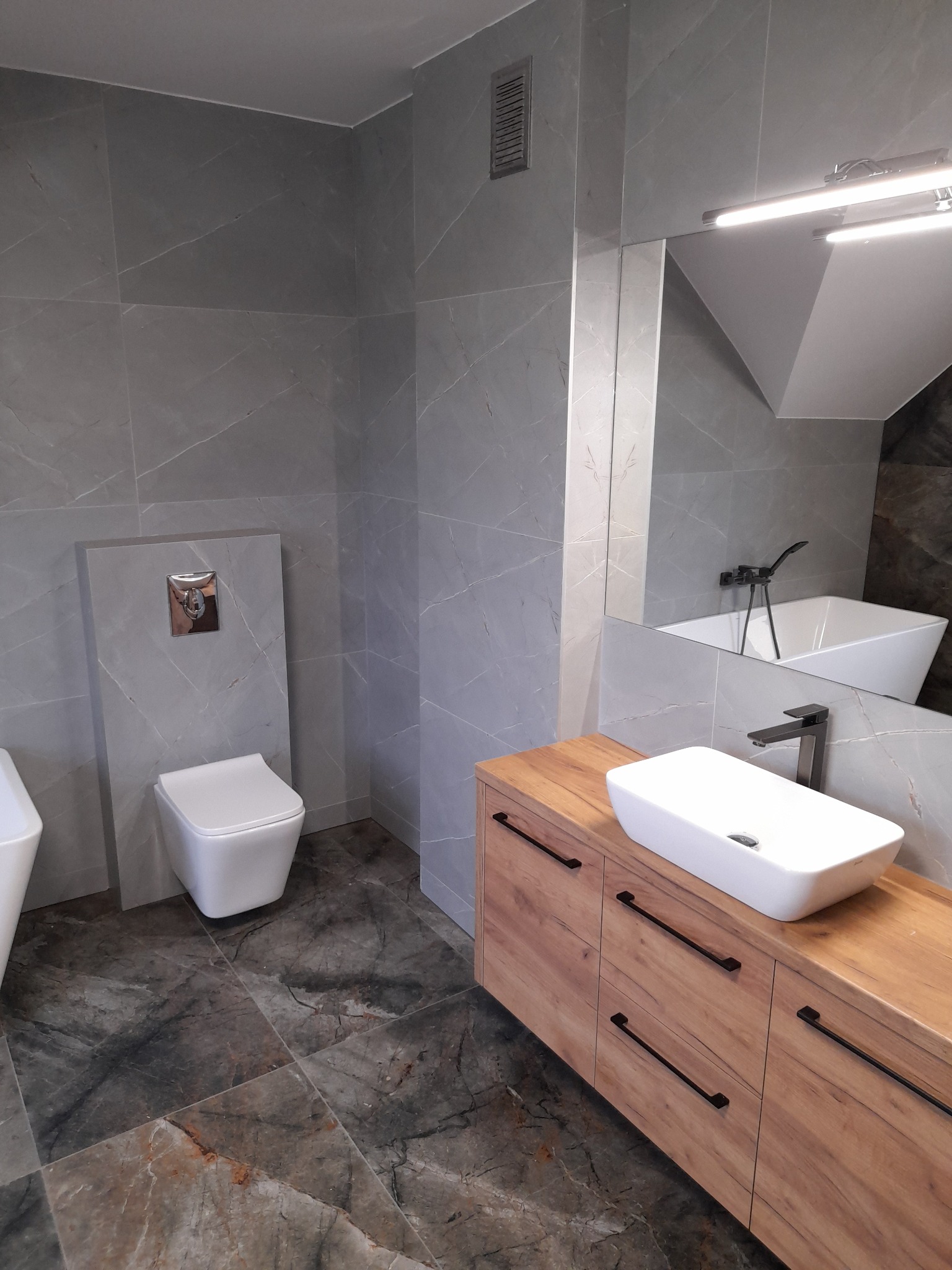 Nowoczesna łazienka z szarymi płytkami imitującymi marmur, drewnianą szafką pod umywalką i wiszącą toaletą. Uwagę przyciąga oryginalna tekstura płytek podłogowych.
