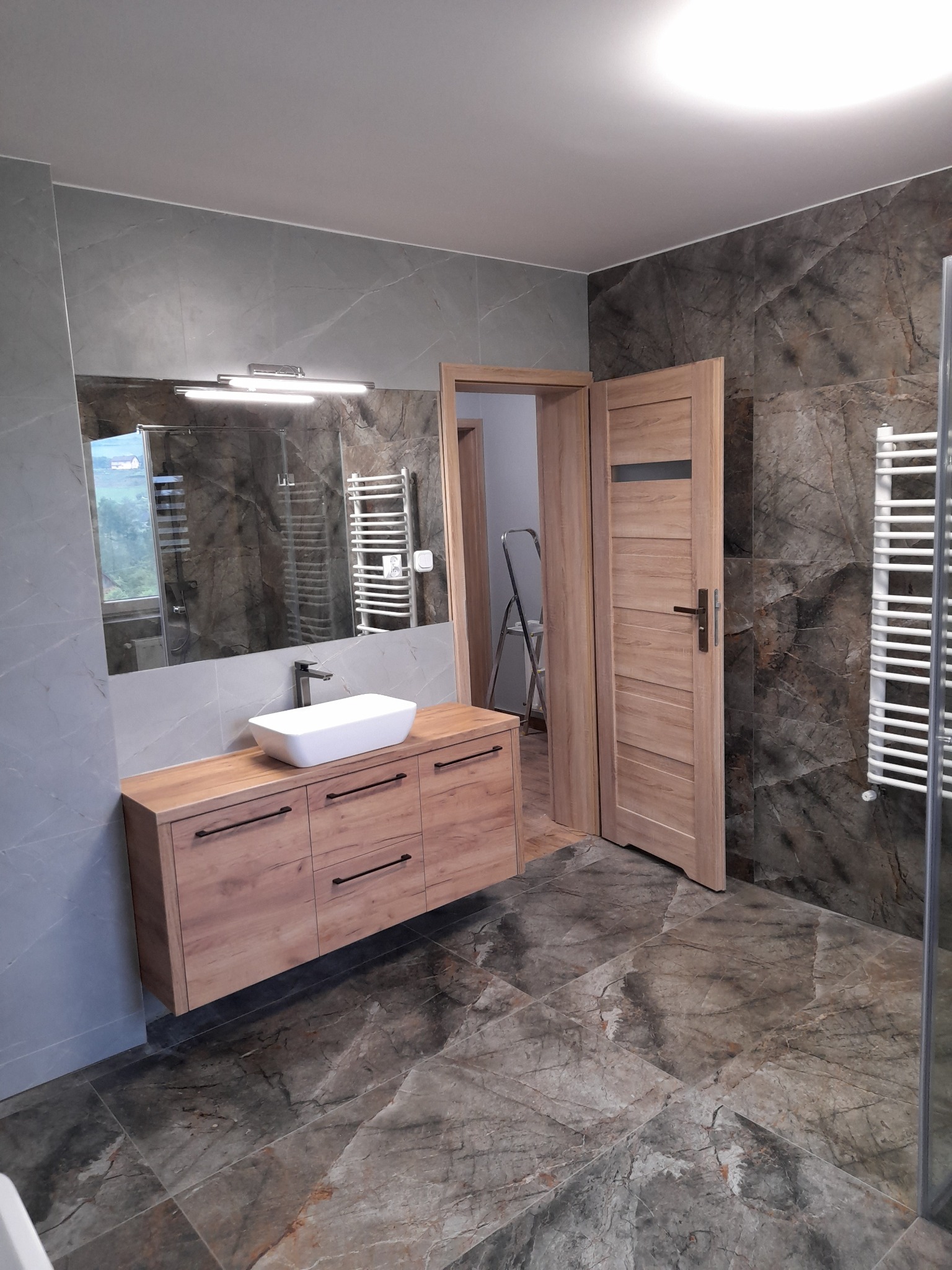 Nowoczesna łazienka z szafką pod umywalką w kolorze drewna i marmurowymi płytkami na ścianach oraz podłodze. Widoczne lustro z oświetleniem i grzejnik drabinkowy.