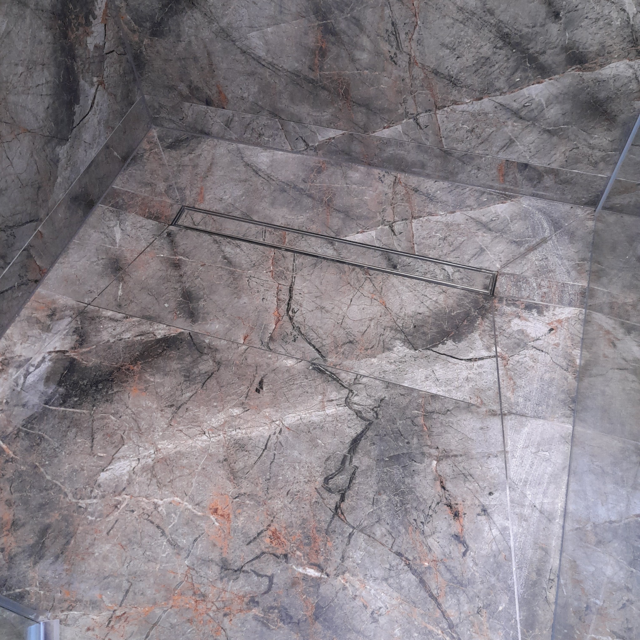 Wykończona kabina prysznicowa z szarymi płytkami imitującymi marmur i odpływem liniowym. Widok z góry, ukazujący precyzję ułożenia płytek i estetykę.