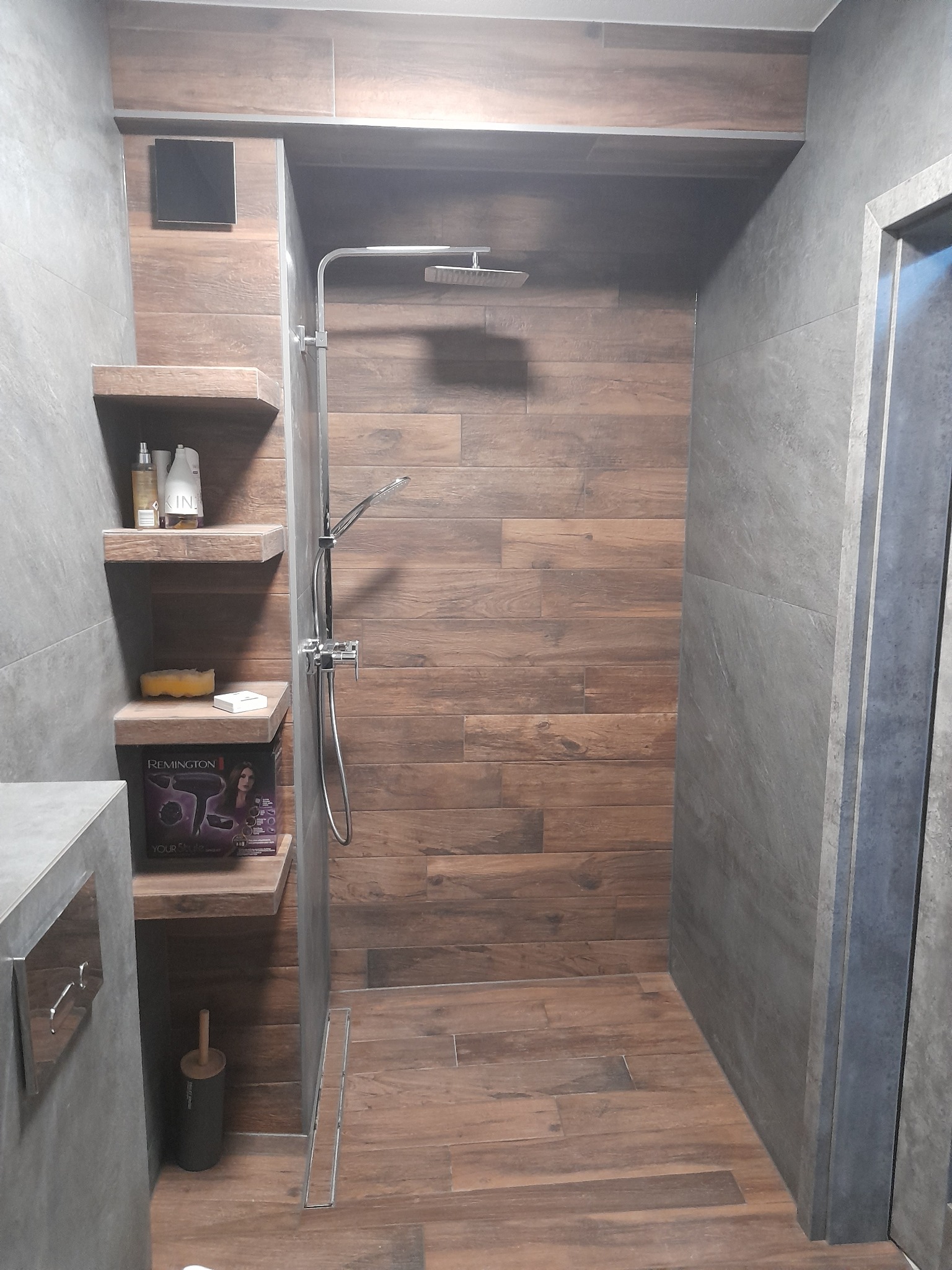 Nowoczesna kabina prysznicowa z drewnopodobnymi płytkami i szarymi ścianami, półki z akcesoriami, minimalistyczny design, widoczny odpływ liniowy.