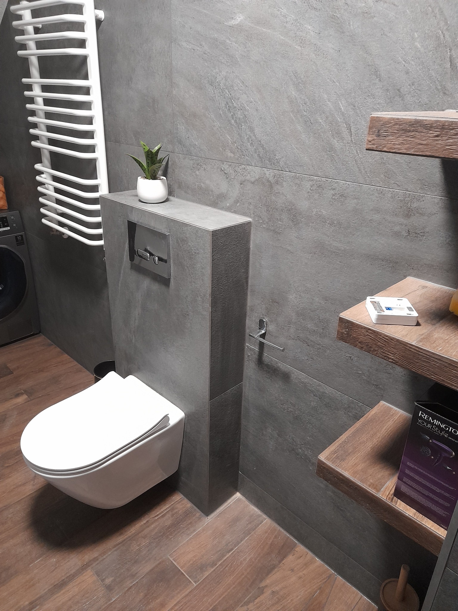 Nowoczesna łazienka z szarymi płytkami, wiszącą toaletą i drewnianymi półkami. Minimalistyczny design z grzejnikiem drabinkowym i akcentem roślinnym.