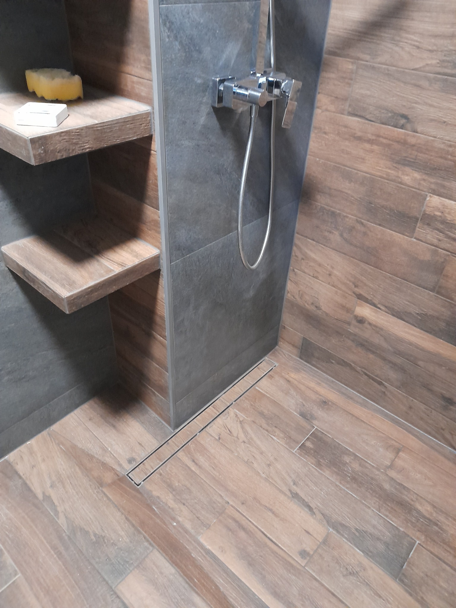 Nowoczesna kabina prysznicowa z drewnopodobnymi płytkami, szarą ścianką i odpływem liniowym. Widoczne półki z akcesoriami i chromowana bateria.