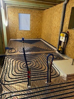 Układ ogrzewania podłogowego z rurami na folii aluminiowej w surowym wnętrzu z płyt OSB, widoczna instalacja pionowa i okno.