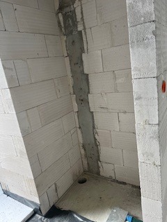 Pionowy kanał instalacyjny w ścianie z białych bloczków, przygotowany do położenia rur. Widoczny otwór w podłodze. Szara zaprawa wypełnia przestrzeń.