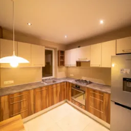 Nowoczesna kuchnia z drewnianymi frontami szafek, beżowymi górnymi szafkami i srebrną lodówką Samsung. Na ścianie dekoracyjny napis w różnych językach.