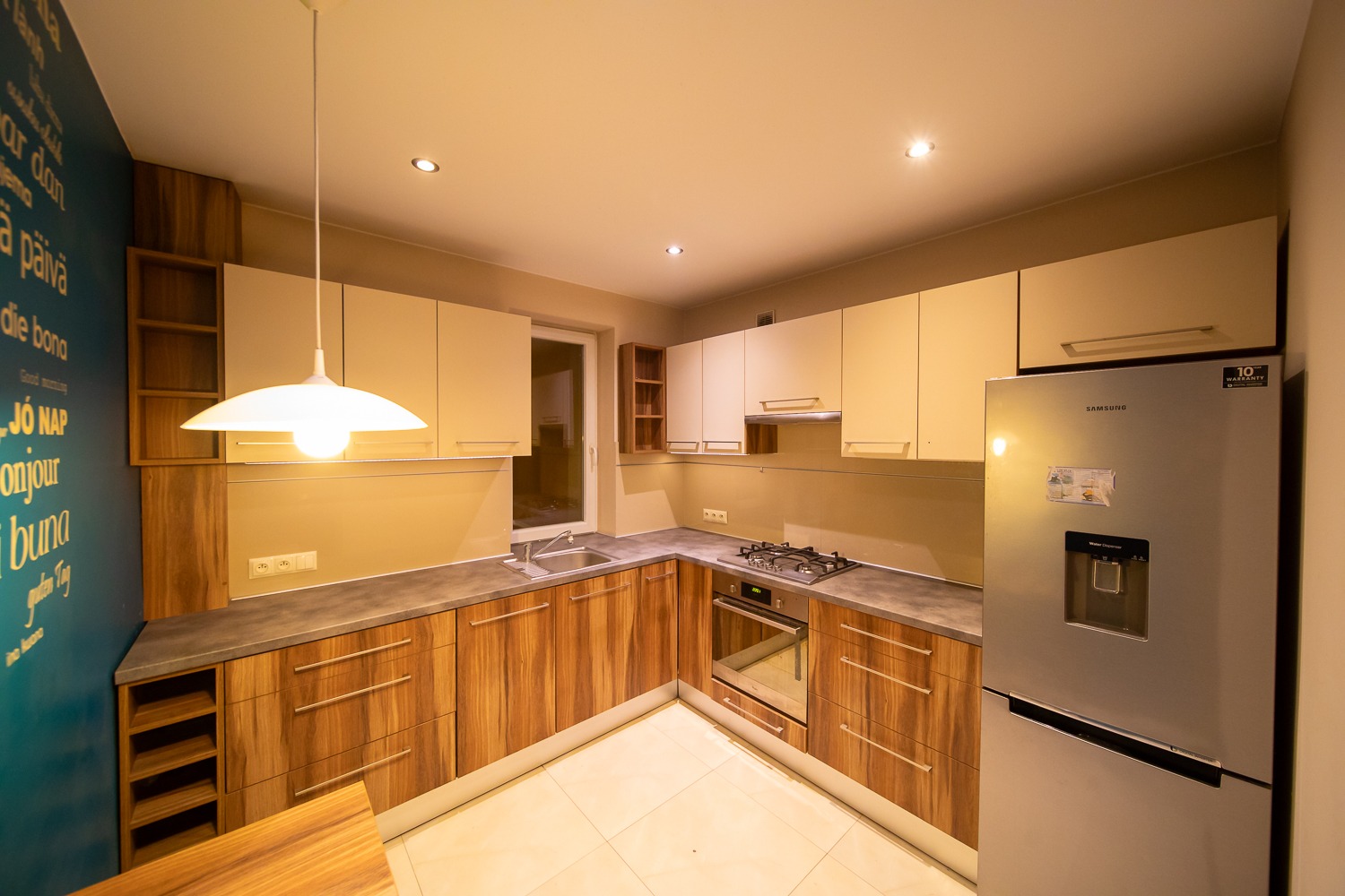Nowoczesna kuchnia z drewnianymi frontami szafek, beżowymi górnymi szafkami i srebrną lodówką Samsung. Na ścianie dekoracyjny napis w różnych językach.