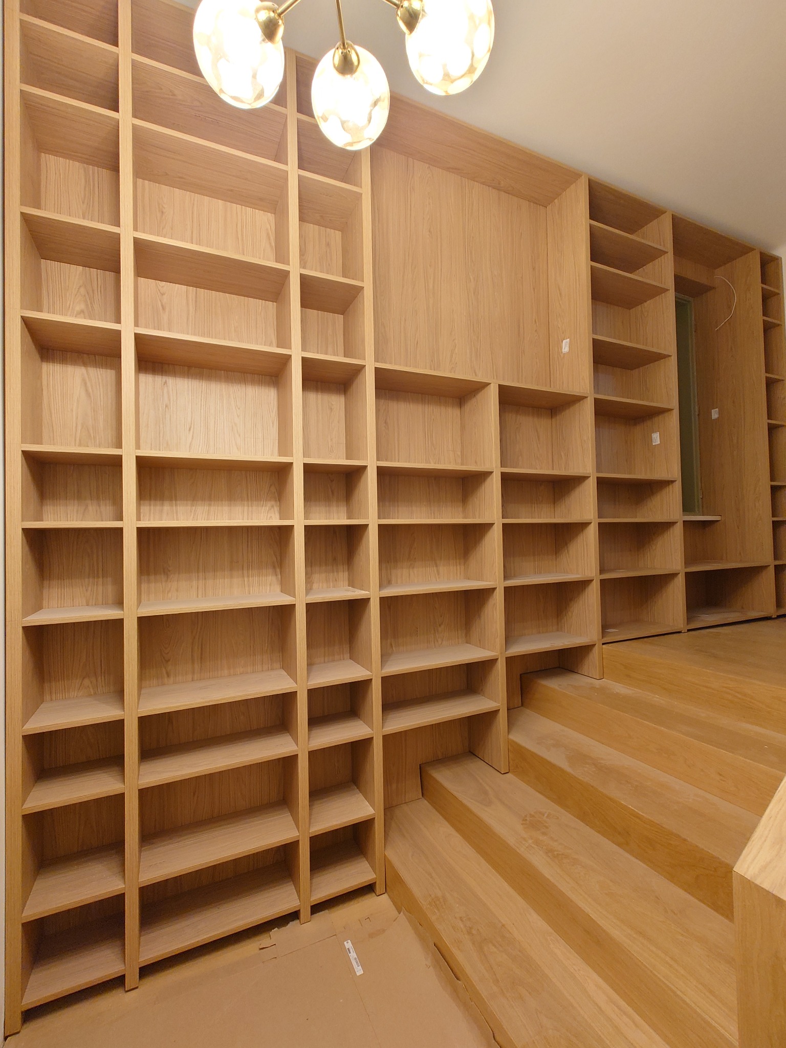 Drewniana zabudowa biblioteczna z półkami, zintegrowana ze schodami. Ciepłe oświetlenie i naturalny kolor drewna. Widok na dużą, otwartą przestrzeń.