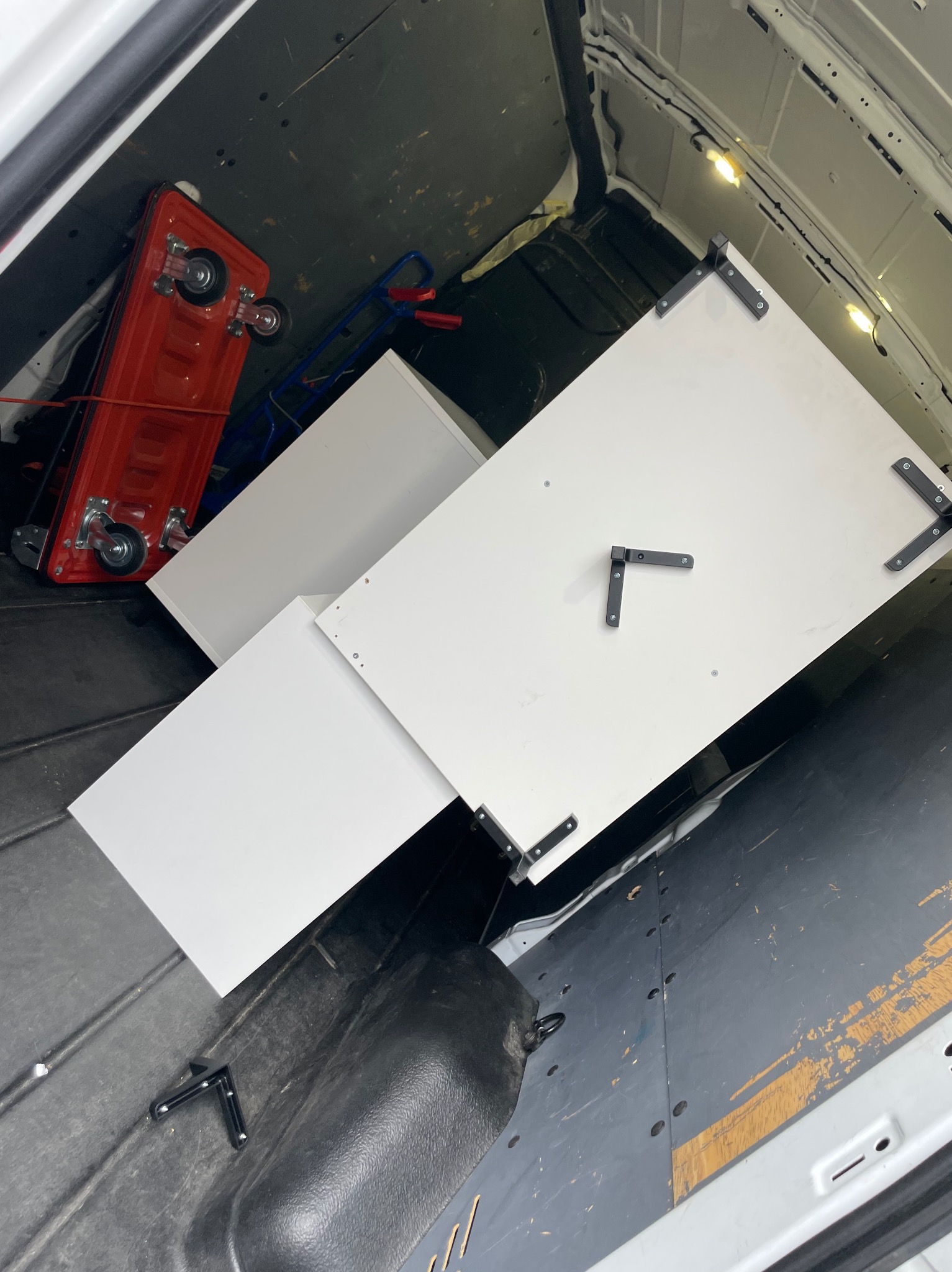 Wnętrze busa z meblami (białe szafki) i wózkiem transportowym. Widok z góry, oświetlenie wewnętrzne. Elementy mocujące na szafkach.