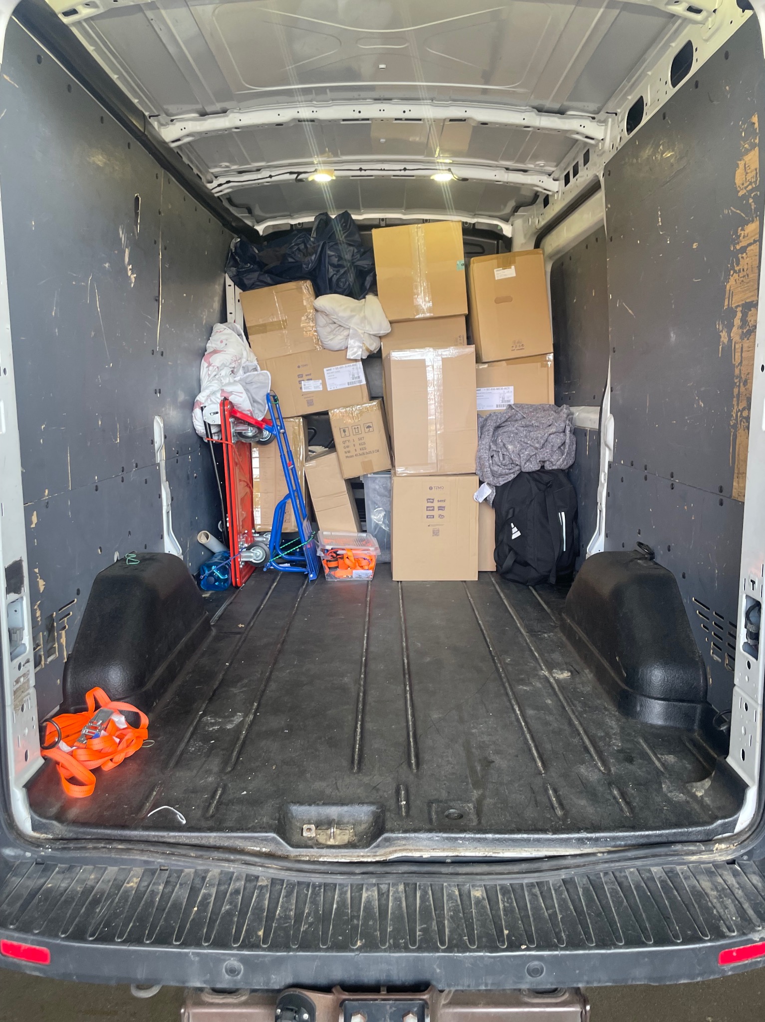 Wnętrze busa wypełnione kartonami, torbami i wózkiem transportowym. Widok z tyłu na częściowo załadowany pojazd przygotowany do przeprowadzki.
