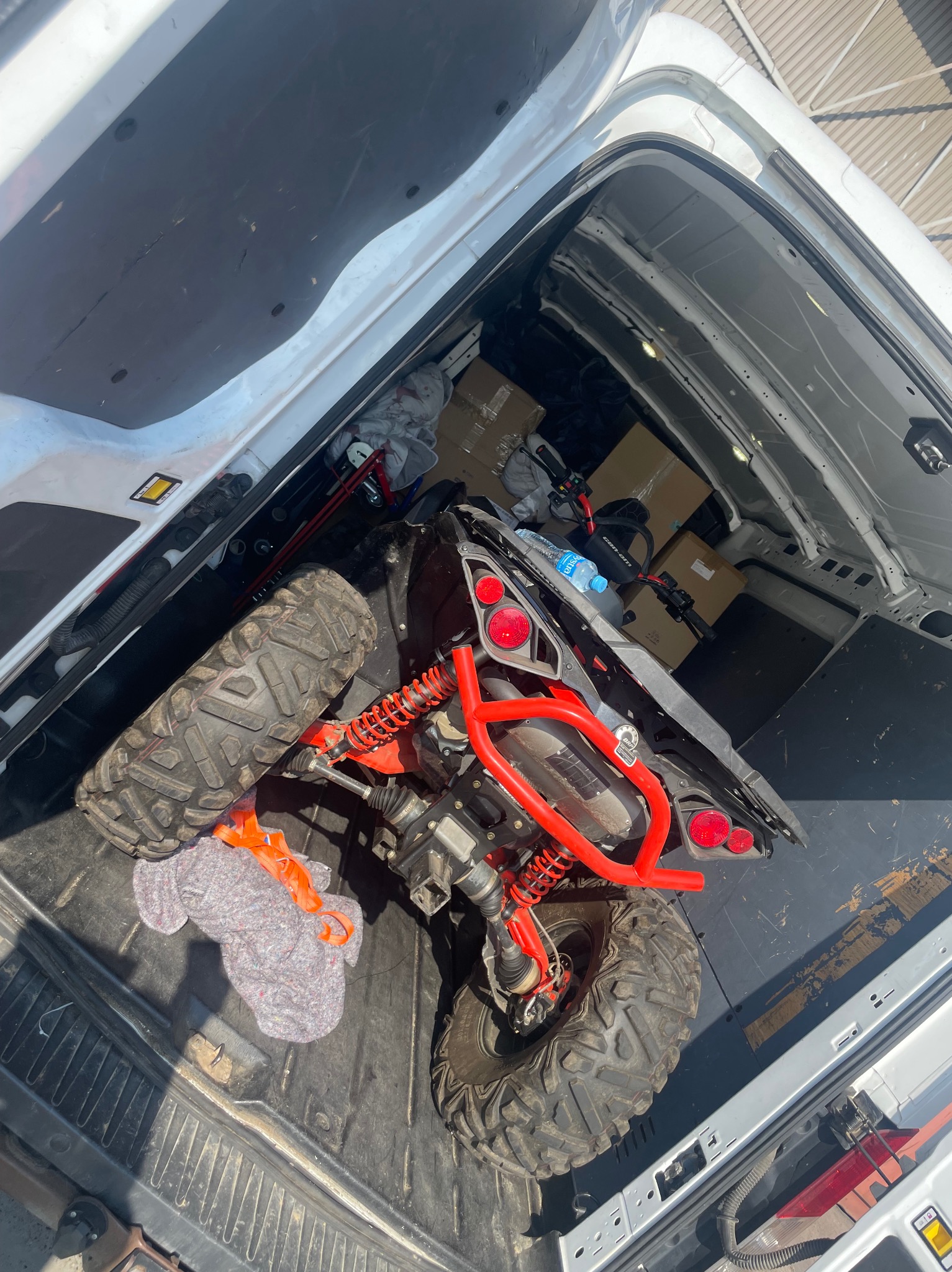 ATV z czerwonymi elementami w przestrzeni ładunkowej białego busa, obok kartony i zabezpieczenia. Widok z góry na załadunek pojazdu.