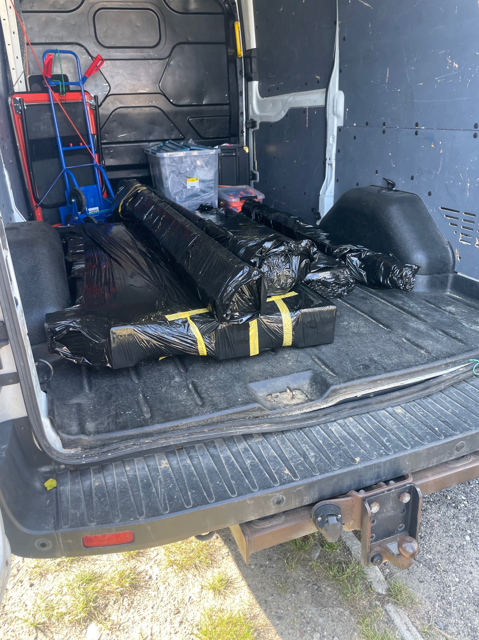 Wnętrze brudnego furgonetki z czarnymi pakunkami zabezpieczonymi taśmą, w tle wózek transportowy i pojemniki. Widoczny hak holowniczy.
