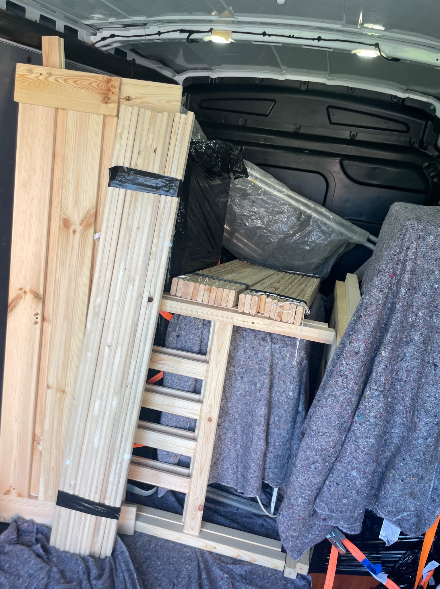 Drewniane elementy mebli zabezpieczone w busie podczas transportu, owinięte kocami i folią dla ochrony przed uszkodzeniami. Widoczne pasy mocujące.