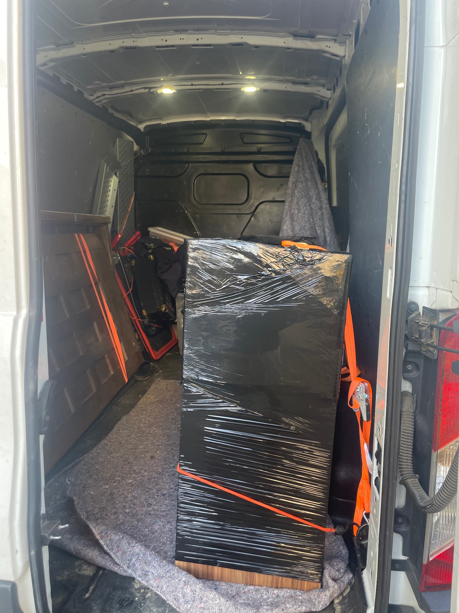 Wnętrze busa transportowego: meble owinięte folią stretch, zabezpieczone pasami transportowymi. Częściowo widoczne drzwi i inne przedmioty. Przygotowanie do transportu.