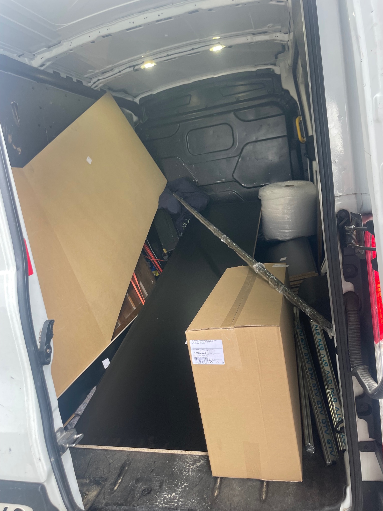 Wnętrze białego busa załadowane kartonami, płytami i folią stretch. Widoczne zabezpieczenia transportowe i elementy wyposażenia przestrzeni ładunkowej.