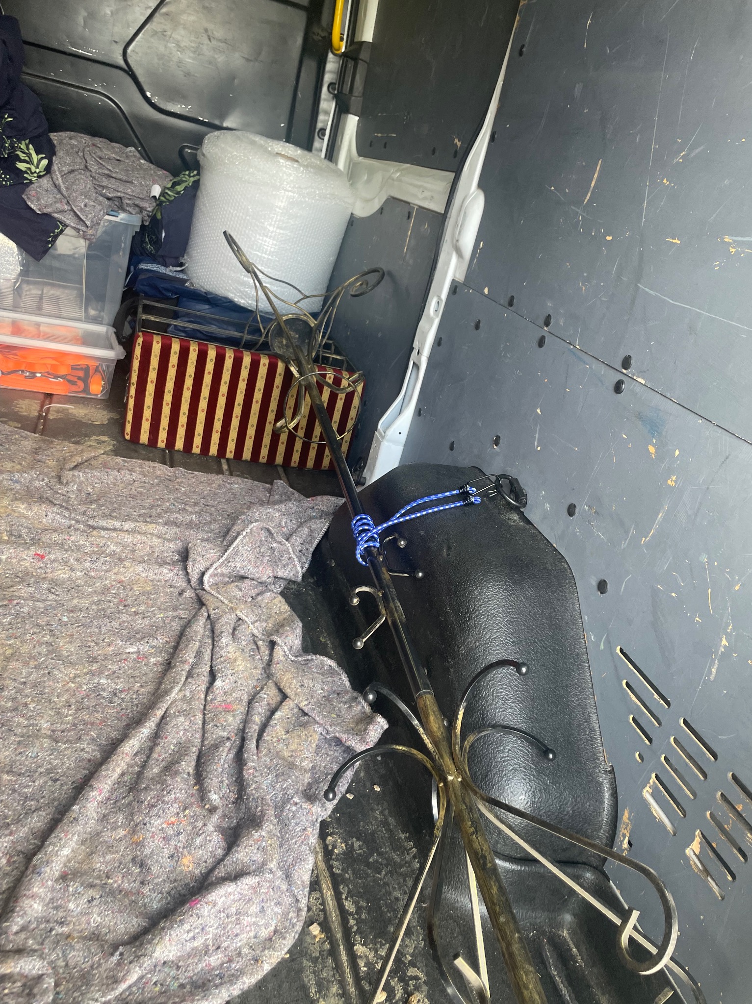 Wnętrze dostawczaka z zabezpieczonym bagażem: pudełko w paski, folia bąbelkowa, ubrania i wieszak na ubrania, przykryte szarą tkaniną.