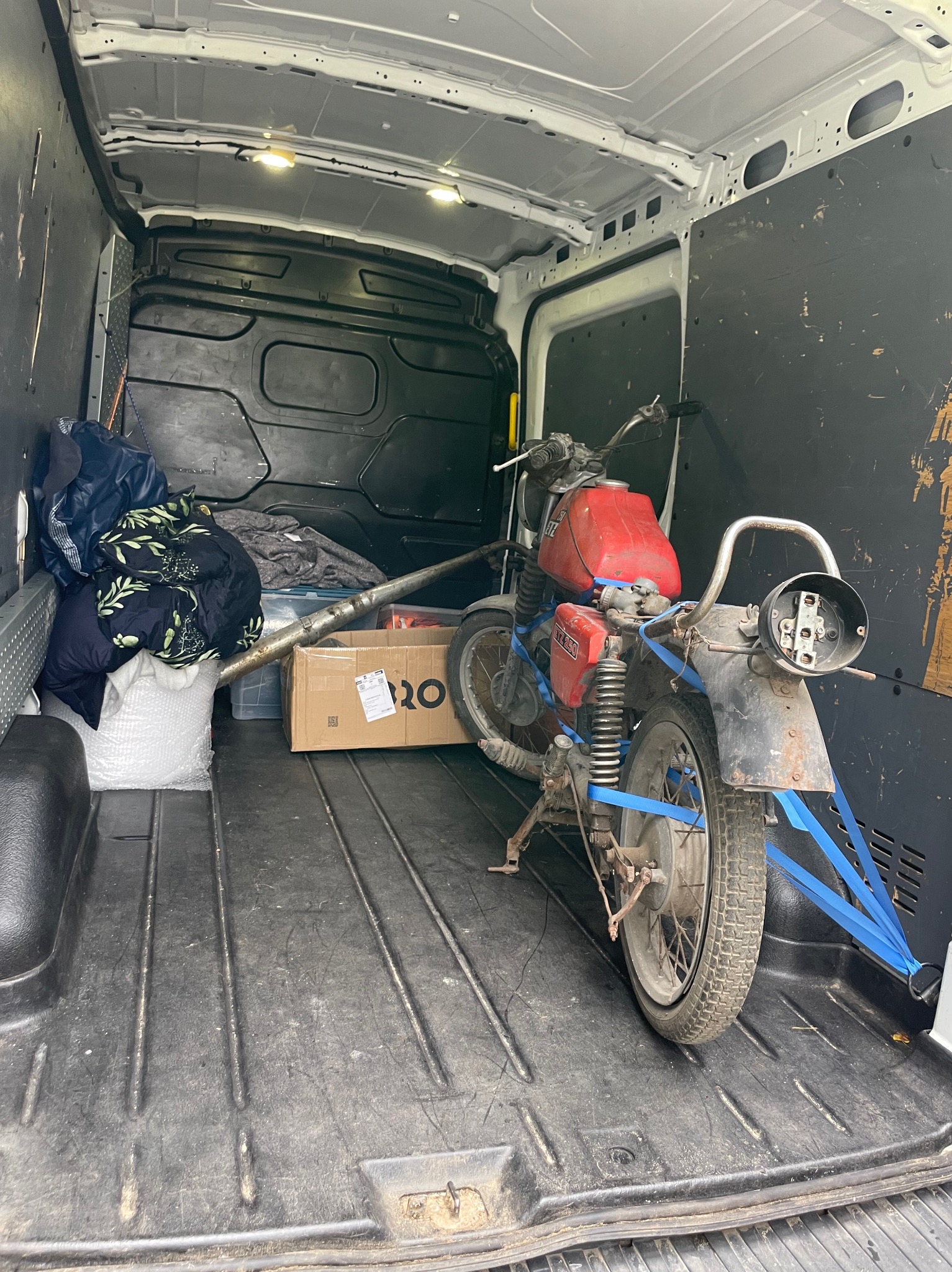 Wnętrze busa z motocyklem ETZ 250 zabezpieczonym niebieskimi pasami, obok karton i worek z odzieżą. Transport motocykli busem.