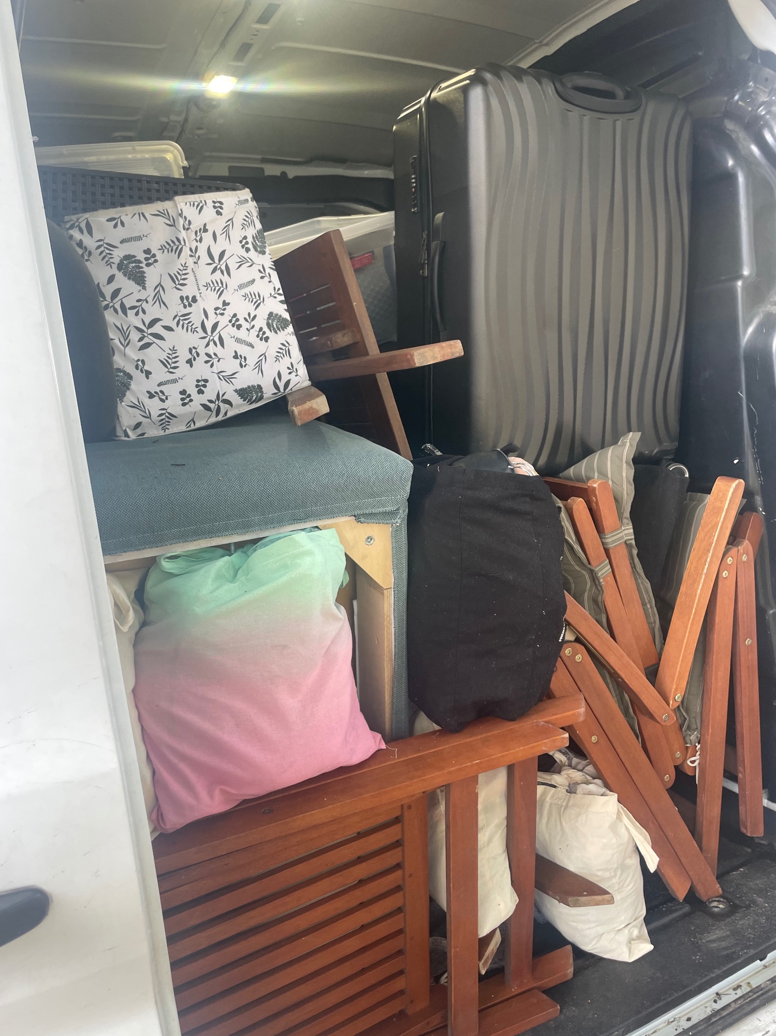 Wnętrze busa załadowane różnorodnymi przedmiotami: walizka, poduszki w poszewkach z motywem roślinnym, krzesła drewniane, torby i tekstylia. Widok z perspektywy otwartych drzwi.