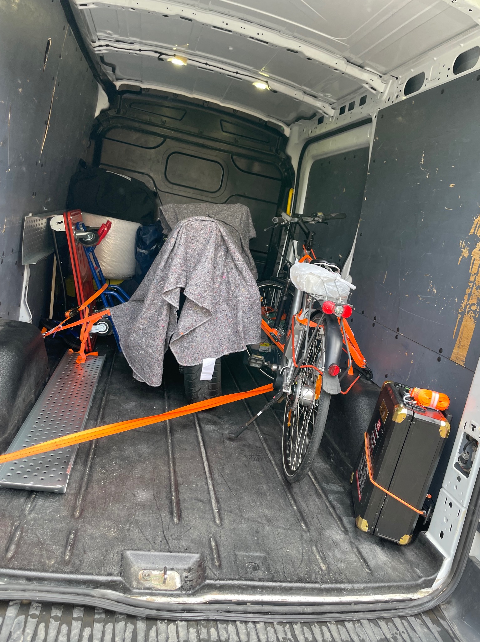 Wnętrze busa z rowerem, wózkiem transportowym i zabezpieczonym bagażem, gotowe do transportu. Widoczne pasy mocujące i koc ochronny.