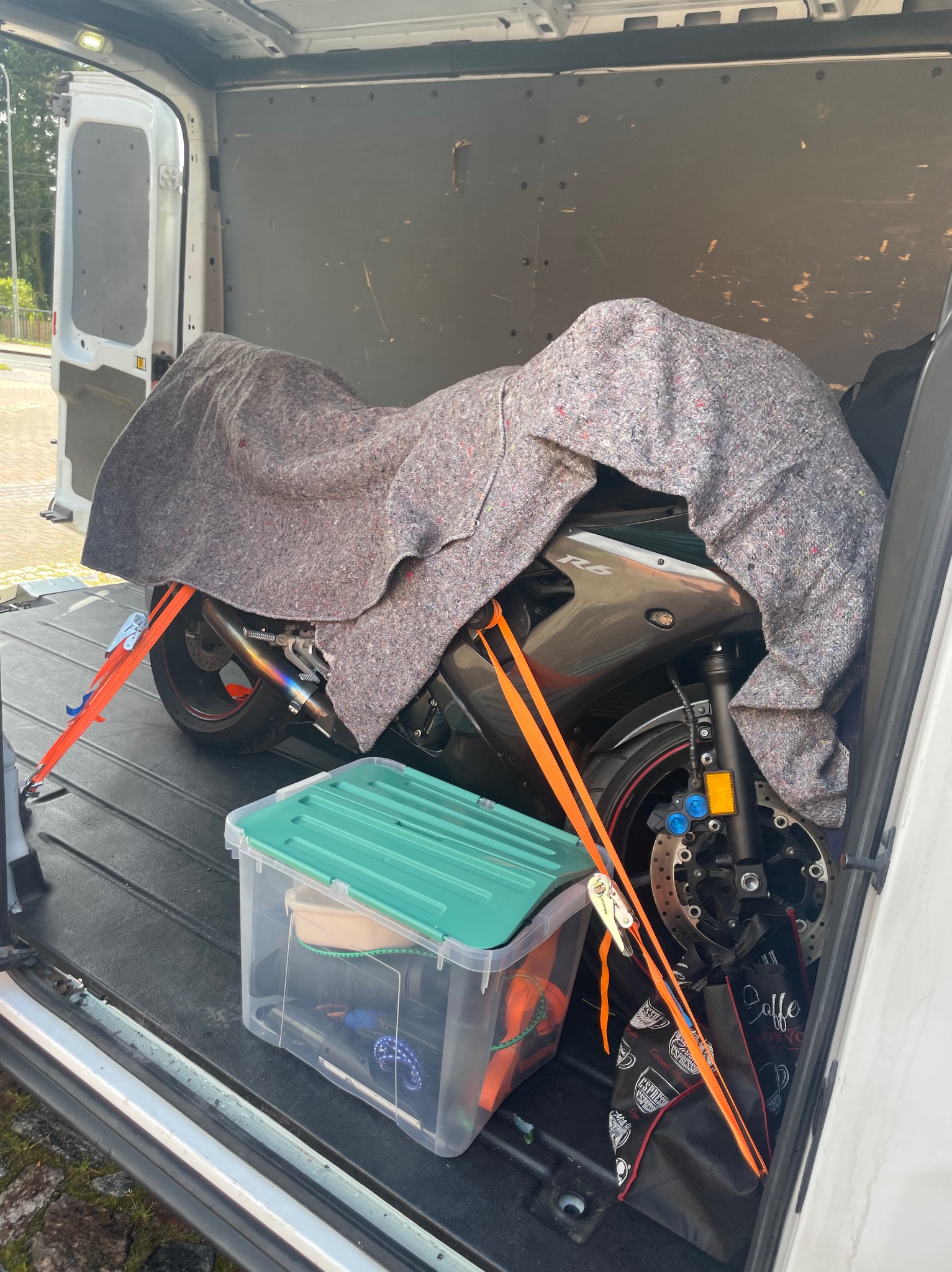 Motocykl R6 zabezpieczony kocem i pasami transportowymi wewnątrz furgonetki, obok przezroczysty pojemnik z akcesoriami. Widok z tyłu pojazdu.