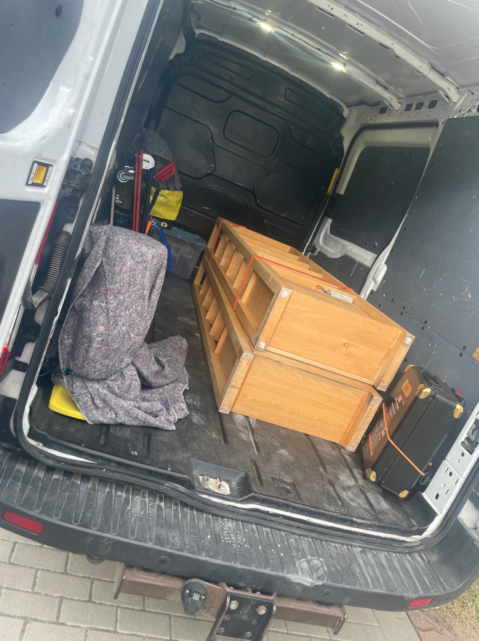 Wnętrze busa transportowego: zabezpieczone meble drewniane, skrzynia narzędziowa i koc ochronny. Widok z tyłu pojazdu, gotowego do transportu.