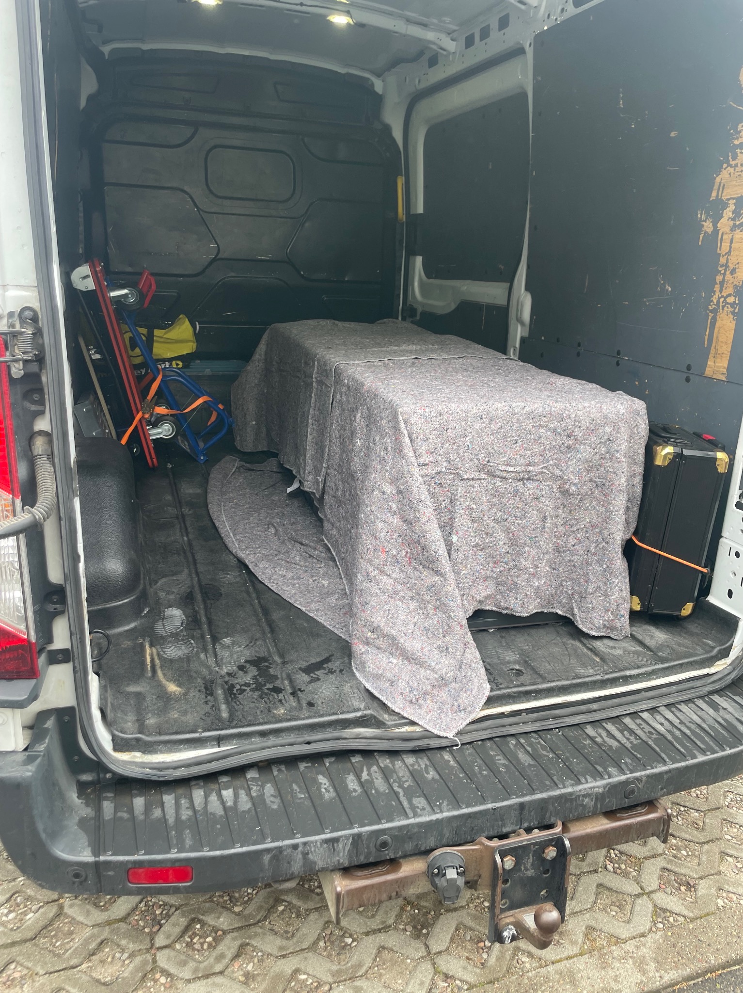 Wnętrze brudnego busa z zabezpieczonym meblem pod szarą narzutą, obok walizka i wózek transportowy. Widok z tyłu pojazdu.