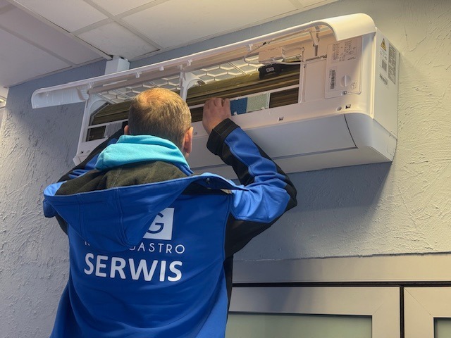 Serwisant w niebieskiej bluzie z napisem 'Serwis' naprawia białą klimatyzację na ścianie, z otwartą obudową i widocznymi filtrami.