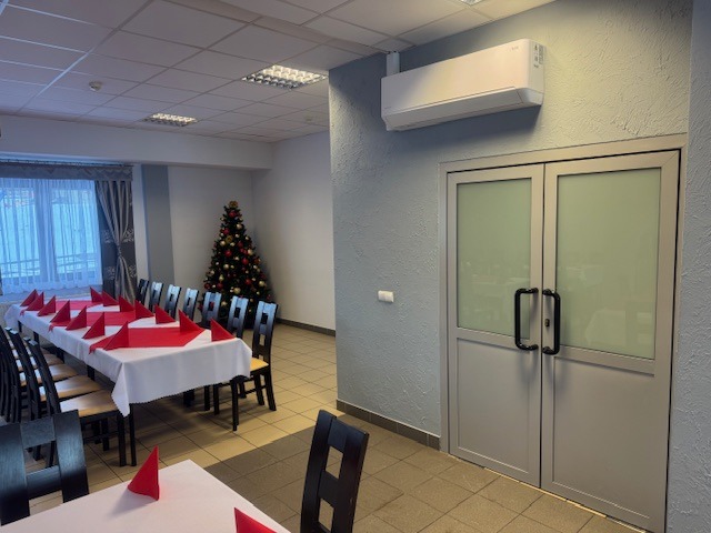 Biała klimatyzacja ścienna nad szarymi drzwiami w sali z nakrytymi stołami i choinką w tle. Widok na salę z klimatyzatorem.