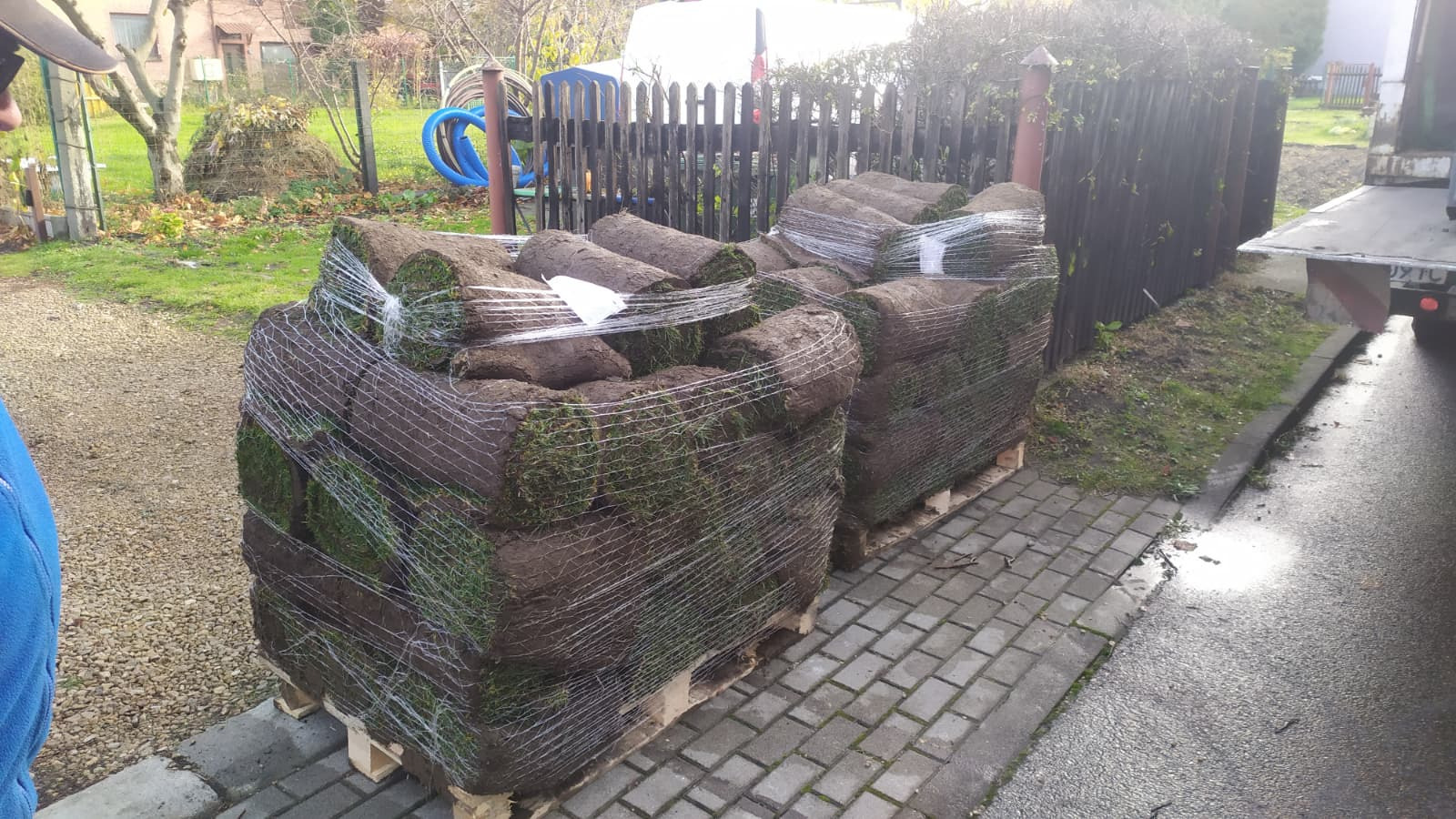 Dwie palety z rolkami trawy, zabezpieczone siatką i folią, stoją na chodniku z kostki brukowej. W tle widoczne ogrodzenie i zieleń.