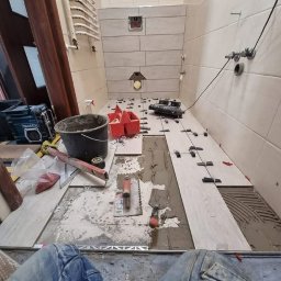 Usługi Budowlane Choszczno - Układanie jasnych płytek w łazience. Widoczny kubeł, narzędzia glazurnicze i fragment ułożonej podłogi. Prace wykończeniowe w toku, z użyciem poziomicy i systemu poziomowania.