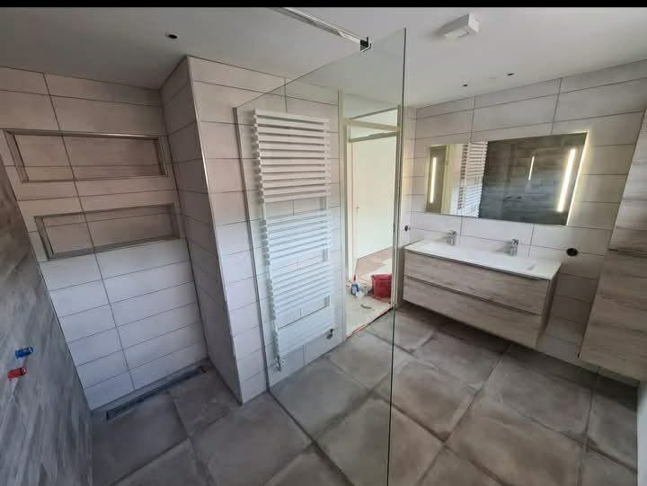 Wykończona łazienka z kabiną prysznicową typu walk-in, grzejnikiem drabinkowym i umywalką z szafką w odcieniach szarości i bieli. Minimalistyczny design z podświetlanym lustrem.