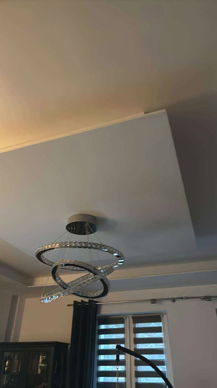 Nowoczesny, kryształowy żyrandol LED w kształcie trzech przeplatających się okręgów, zamontowany na białym, podwieszanym suficie w pokoju z oknem.