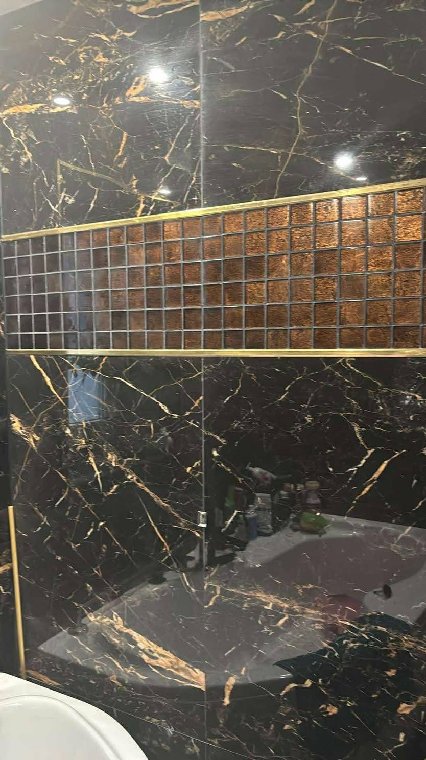 Czarne płytki imitujące marmur z złotymi żyłkami, oddzielone mozaiką z brązowych kwadratów i złotą listwą w wykończonej łazience. Widoczna umywalka i detale wyposażenia.