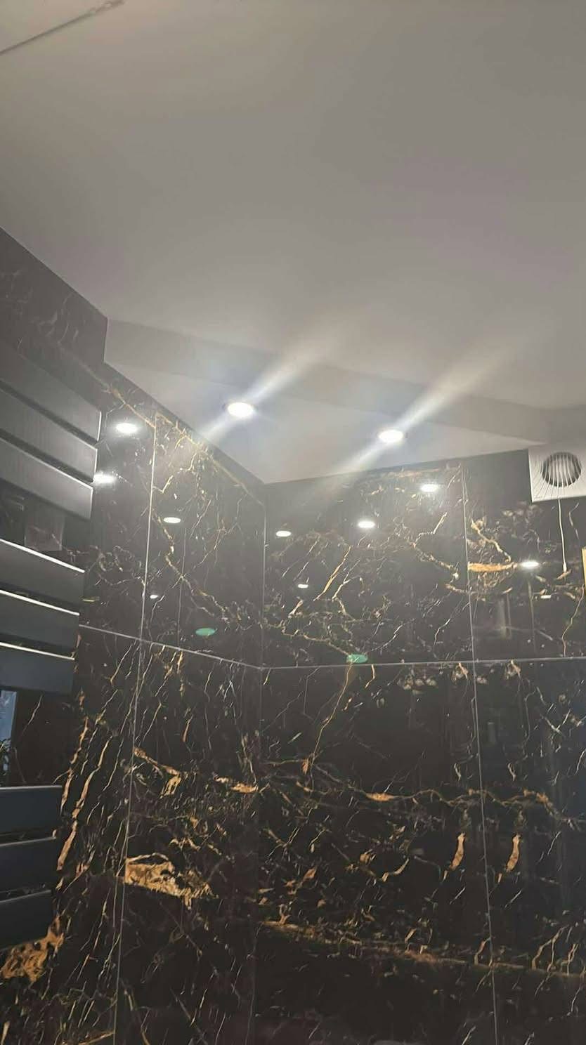 Elegancka łazienka z czarnymi płytkami imitującymi marmur ze złotymi żyłami, oświetlona punktowymi lampami sufitowymi i wyposażona w grzejnik drabinkowy.