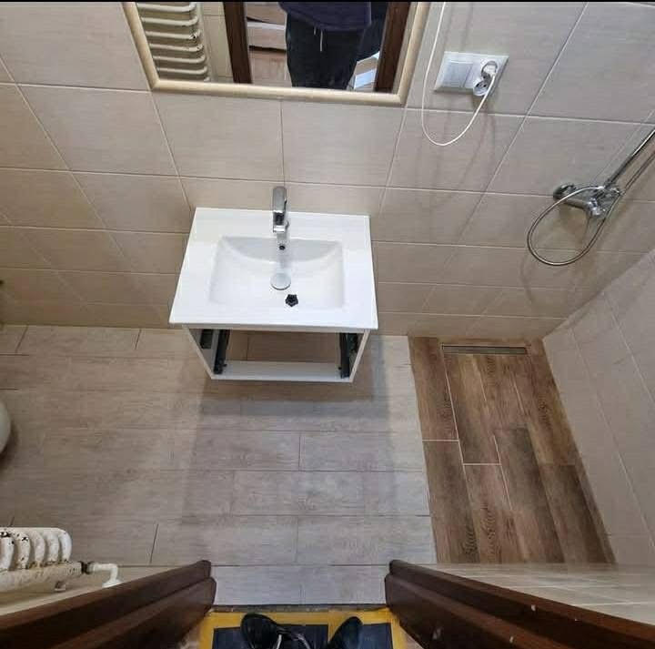 Nowoczesna, jasna łazienka z białą umywalką na tle beżowych płytek i drewnopodobnej podłogi. Widoczny fragment kabiny prysznicowej i lustro z odbiciem.