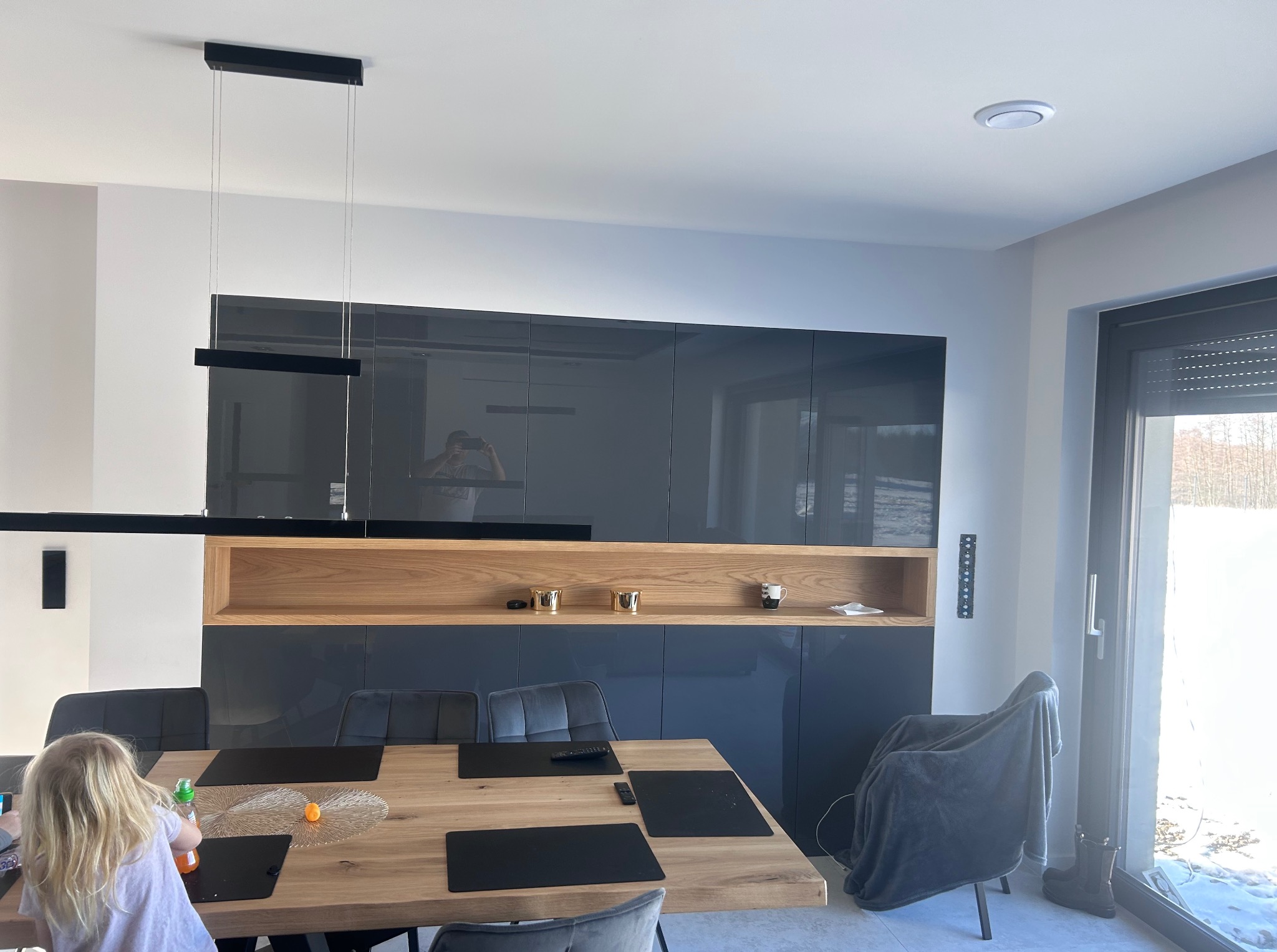 Nowoczesna kuchnia z grafitowymi szafkami i drewnianą wnęką, oświetlona wiszącą lampą. Widoczny stół z krzesłami i fragment osoby. Za oknem zimowy krajobraz.