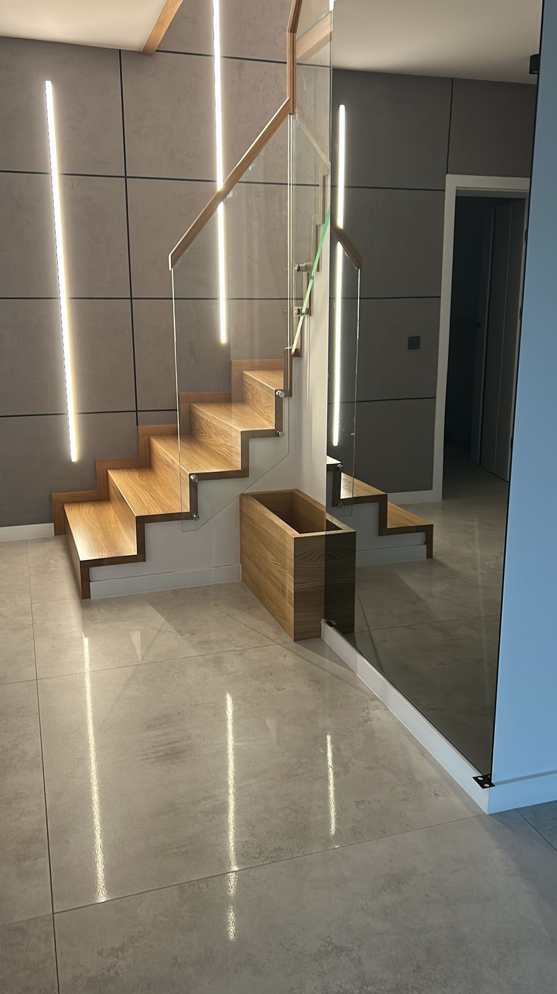 Nowoczesne schody drewniane z szklaną balustradą, podświetlane pionowymi lampami LED, z drewnianą donicą u podstawy. Minimalistyczny design w jasnej kolorystyce.