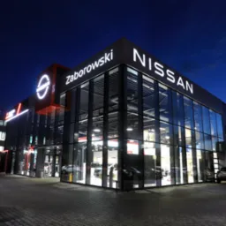 Oświetlony salon samochodowy Nissan Zaborowski nocą; widoczne logo i nazwa dealera. Wnętrze salonu z wystawionymi pojazdami. Na zewnątrz zaparkowane auta.