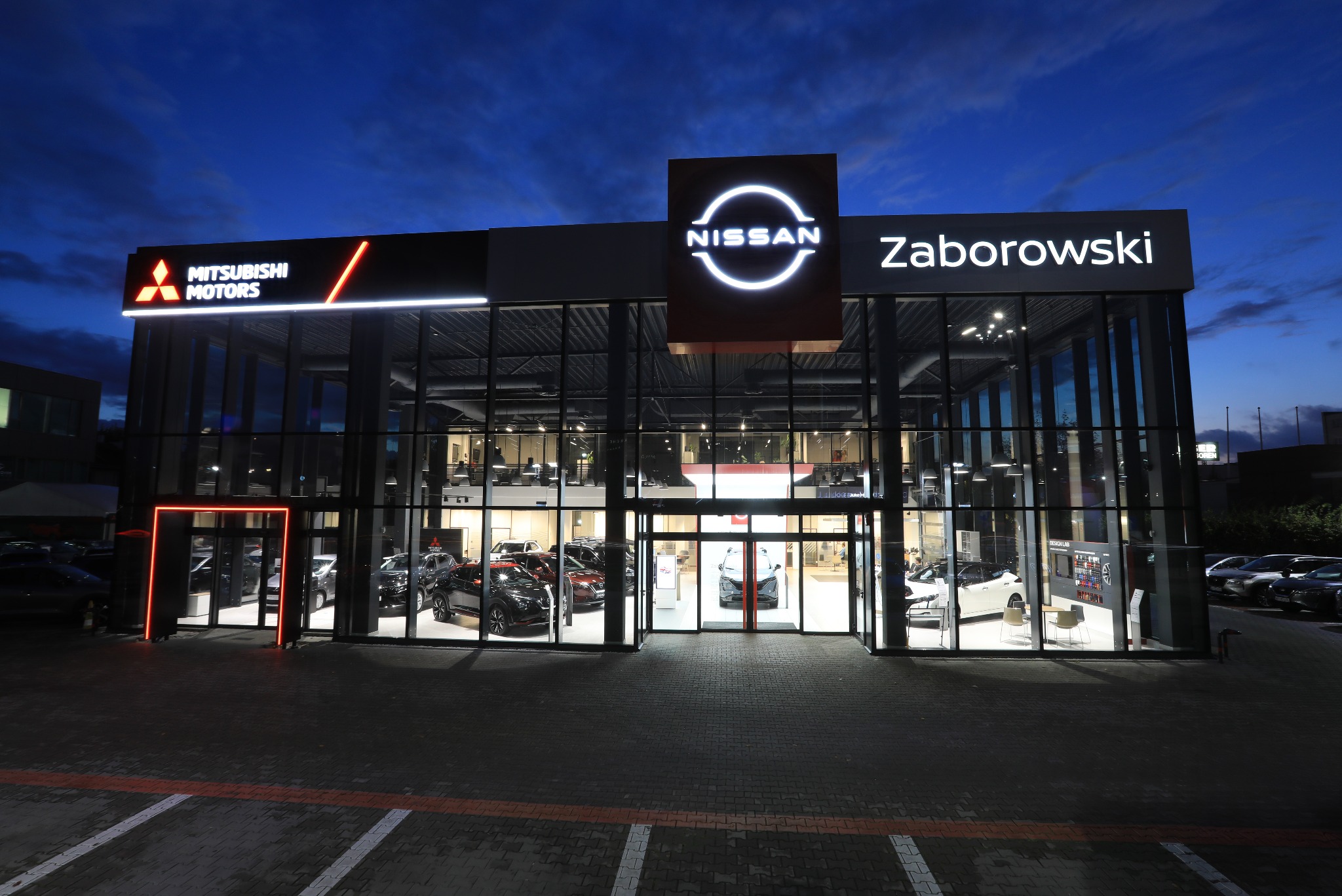 Nowoczesny salon samochodowy Mitsubishi i Nissan z przeszkloną fasadą oświetloną nocą, prezentujący różne modele aut wewnątrz i na zewnątrz budynku.