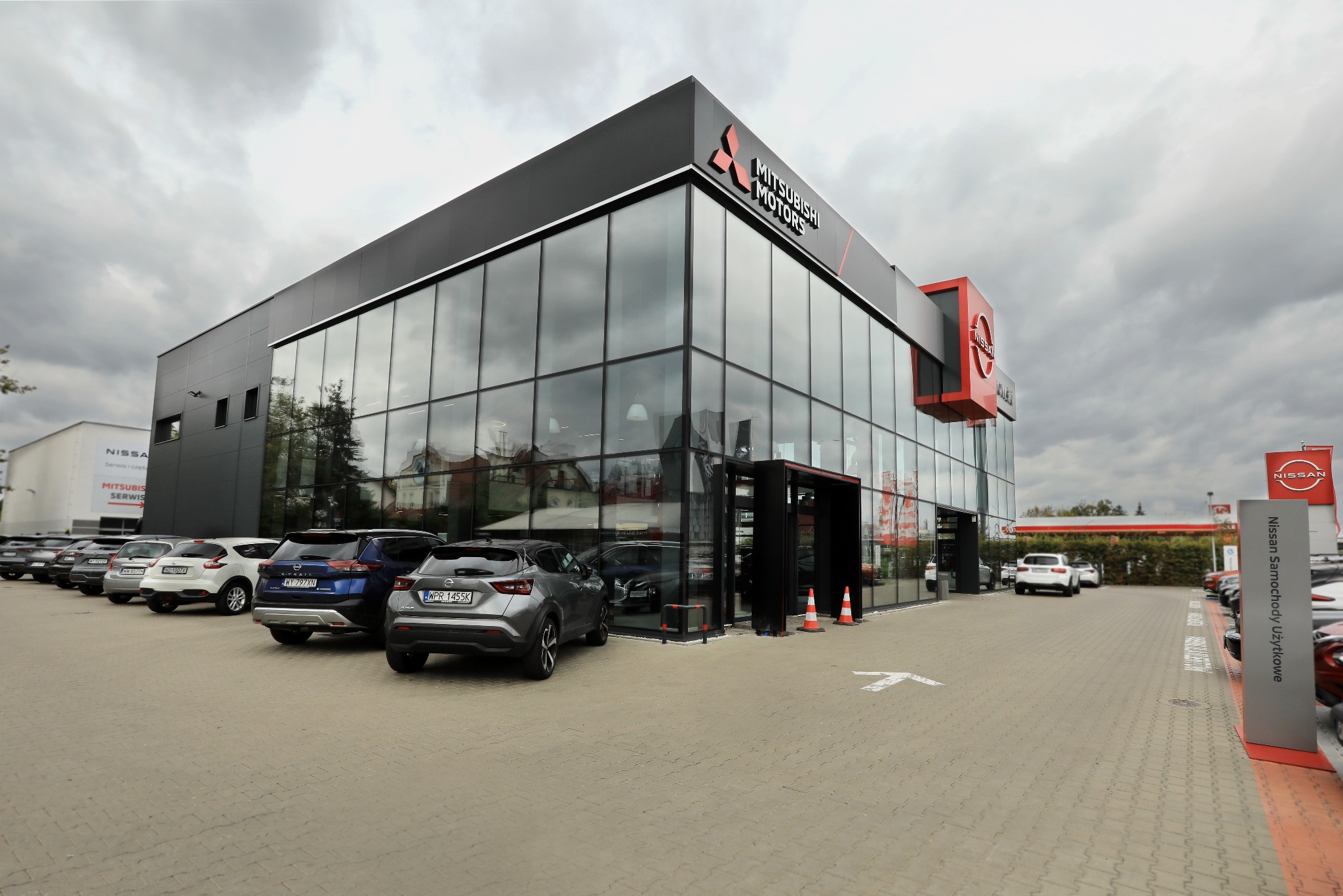 Salon samochodowy Mitsubishi Motors z przeszkloną fasadą i zaparkowanymi pojazdami na zewnątrz. Widoczny również znak Nissan. Pochmurne niebo w tle.