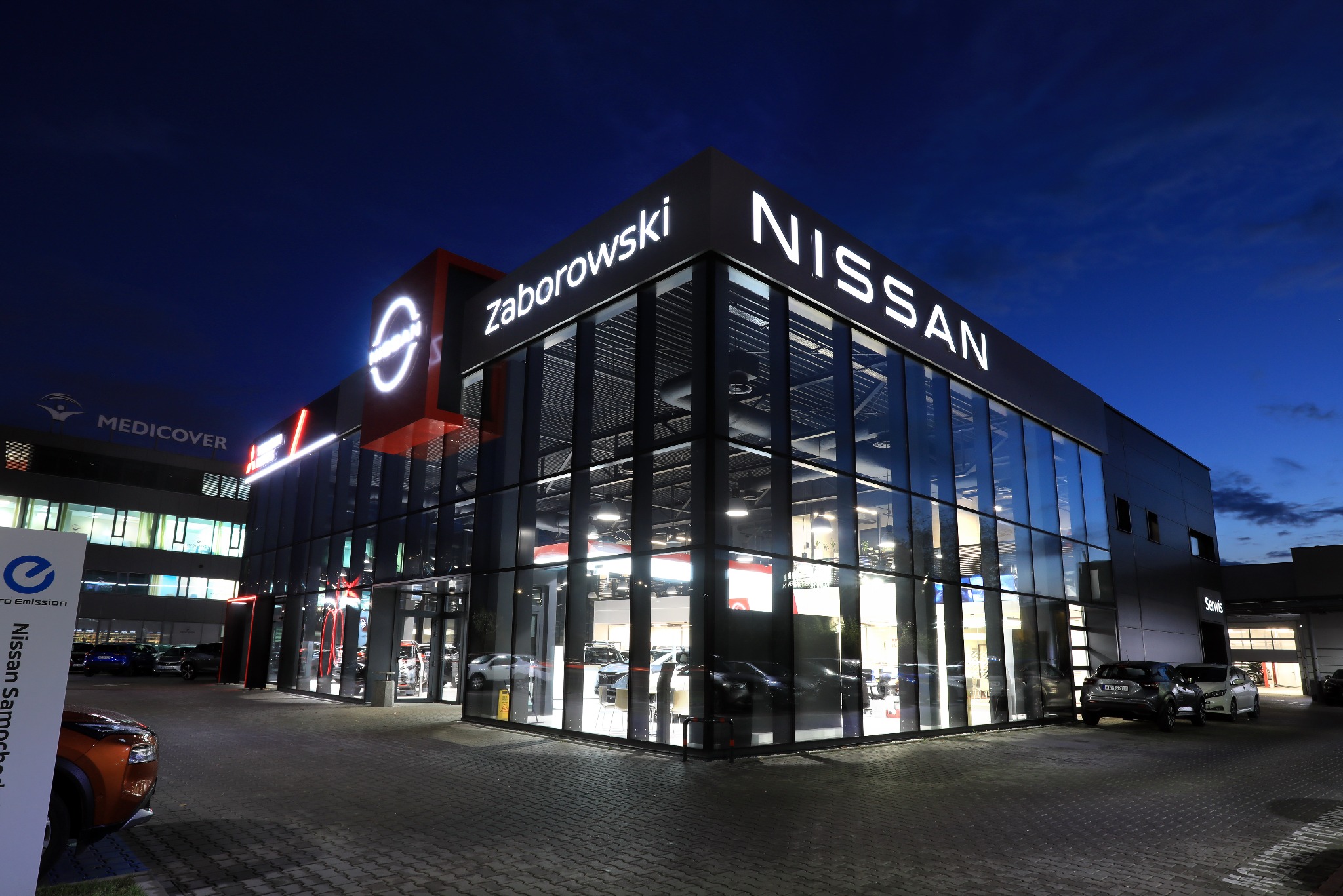 Oświetlony salon samochodowy Nissan Zaborowski nocą; widoczne logo i nazwa dealera. Wnętrze salonu z wystawionymi pojazdami. Na zewnątrz zaparkowane auta.