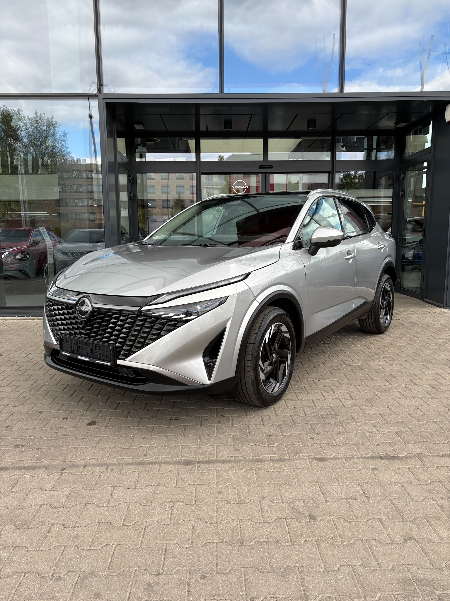 Srebrny Nissan Qashqai zaparkowany przed salonem z przeszkloną fasadą. Widoczne detale karoserii i felgi. Refleksy światła na lakierze, kostka brukowa w tle.