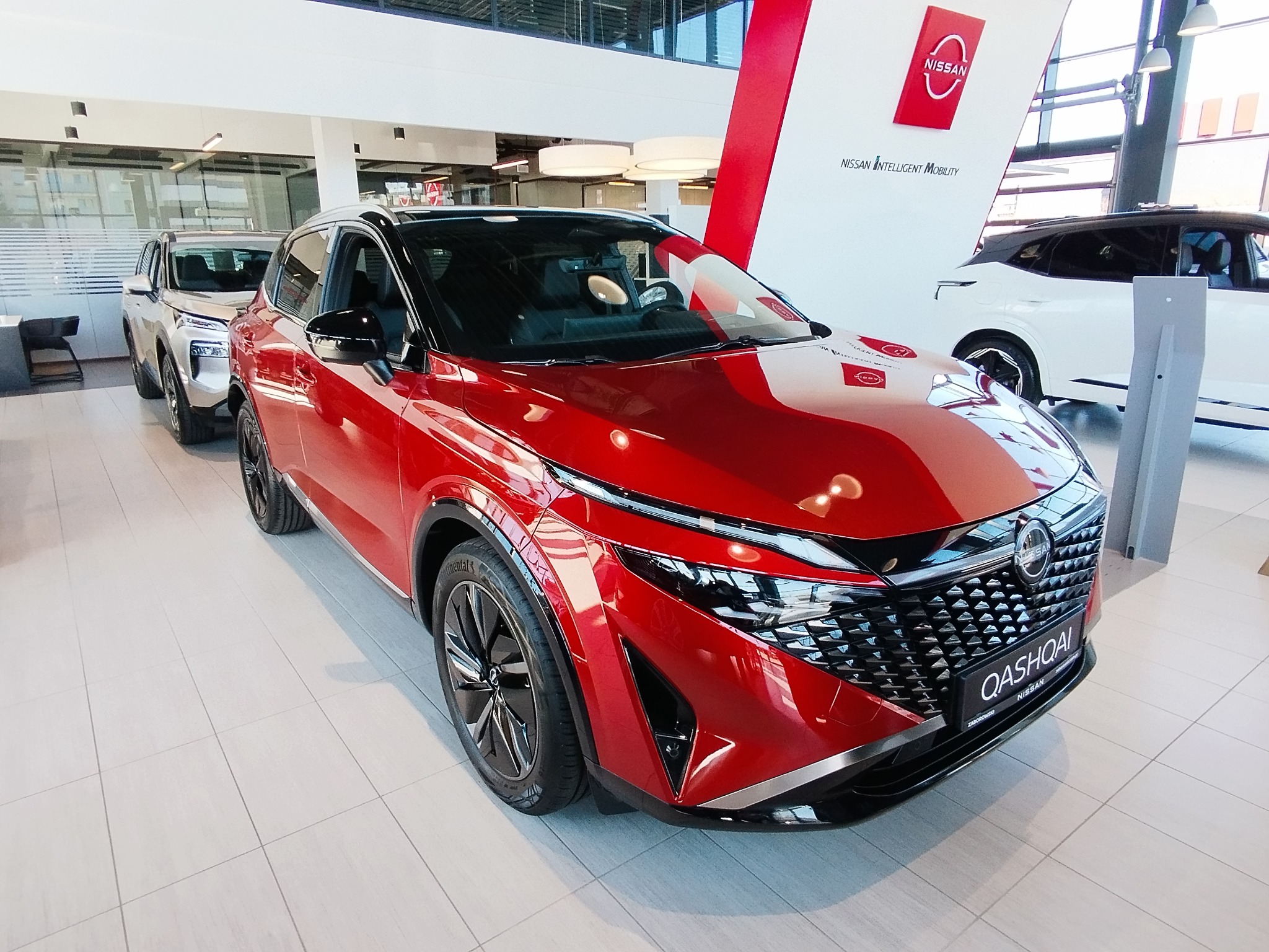Czerwony Nissan Qashqai w salonie samochodowym, widok z przodu, na pierwszym planie felgi i reflektory. W tle inne modele i logo Nissan.