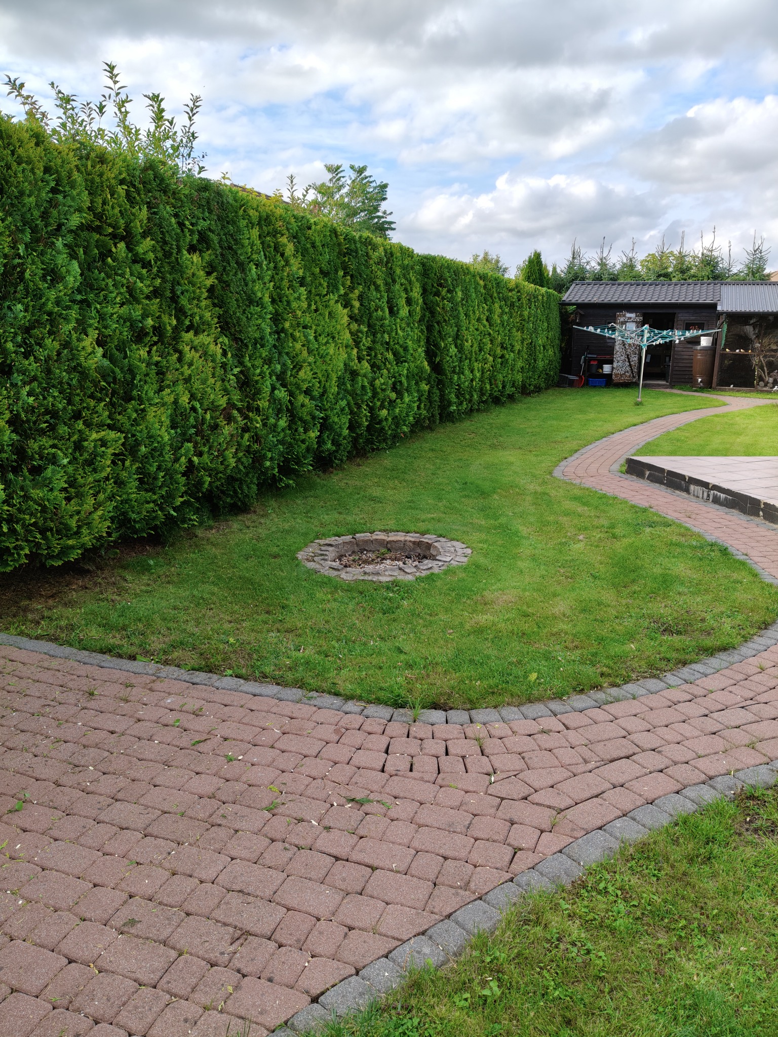 Zadbany ogród z równo przystrzyżonym żywopłotem i ścieżką z kostki brukowej. W tle widoczna altana i suszarka na pranie. Centralnie palenisko.